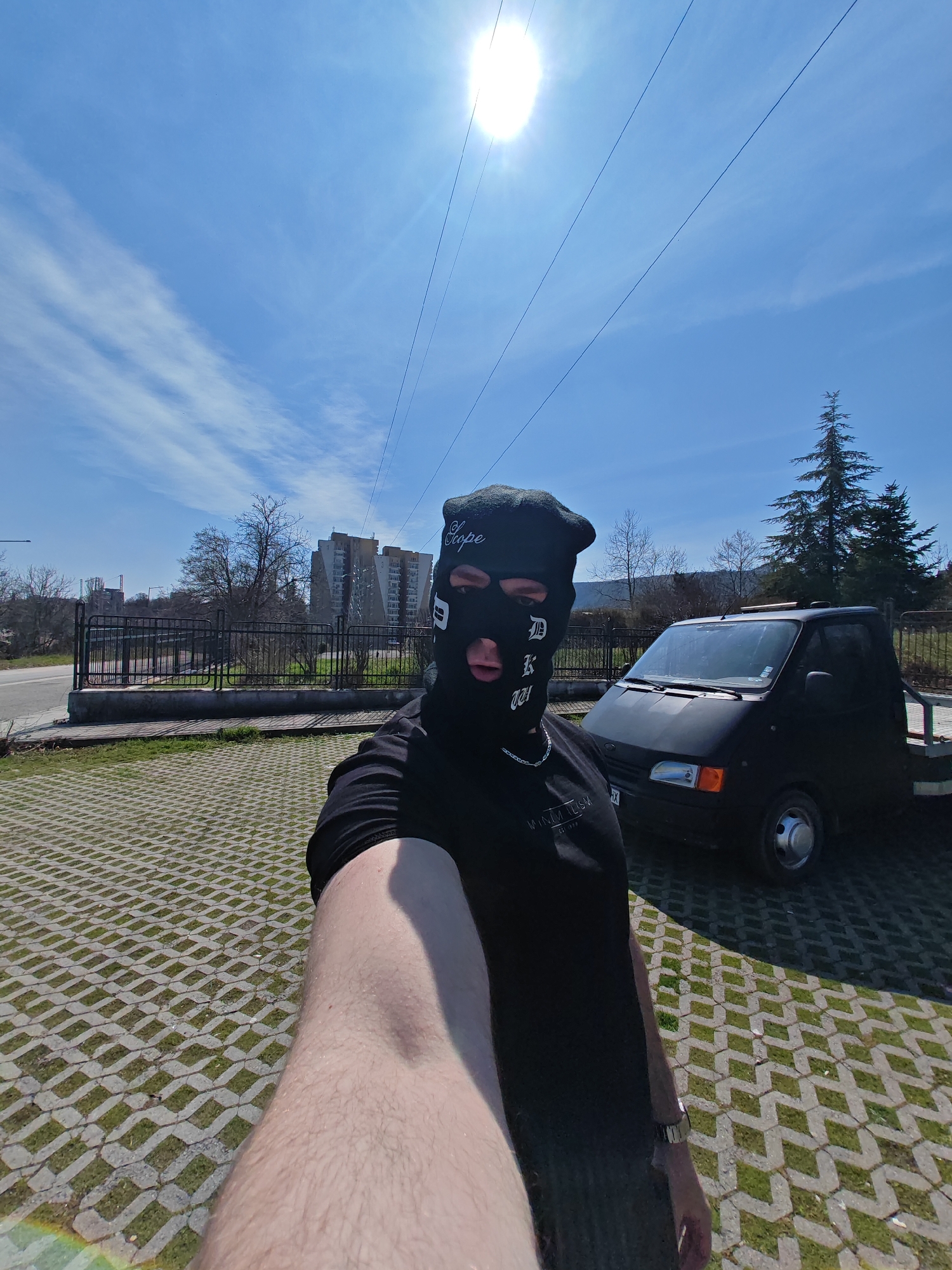The image portrays an outdoor, daylight scene in Shumen, Bulgaria. A single individual, seen from the chest up, is captured in a selfie, holding the camera with their right arm. This individual wears a black balaclava featuring white text: "Kope" on the forehead and "D K 001" below the right eye opening. They are attired in a black short-sleeved t-shirt with partial white text visible and a silver-colored chain necklace. A silver-toned watch is worn on the left wrist.
The subject stands on permeable paving, characterized by grey concrete pavers forming a hexagonal pattern interspersed with grass. Behind the individual, a black metal fence delineates the paved area from a road. Across this road, bare deciduous trees are visible, and in the background, several multi-story residential buildings, consistent with apartment blocks, are present.
To the subject's right, a black utility vehicle, resembling a modified Ford Transit flatbed or car carrier, is parked. A Bulgarian license plate beginning with "H," indicating Shumen registration, is partially visible on the vehicle's front. A tall, dark green coniferous tree stands behind the vehicle. The sky is clear blue with scattered cirrus clouds. The sun is high in the sky, positioned in the upper right, generating strong light and a lens flare in the lower left foreground. Three overhead power lines traverse the upper portion of the frame.