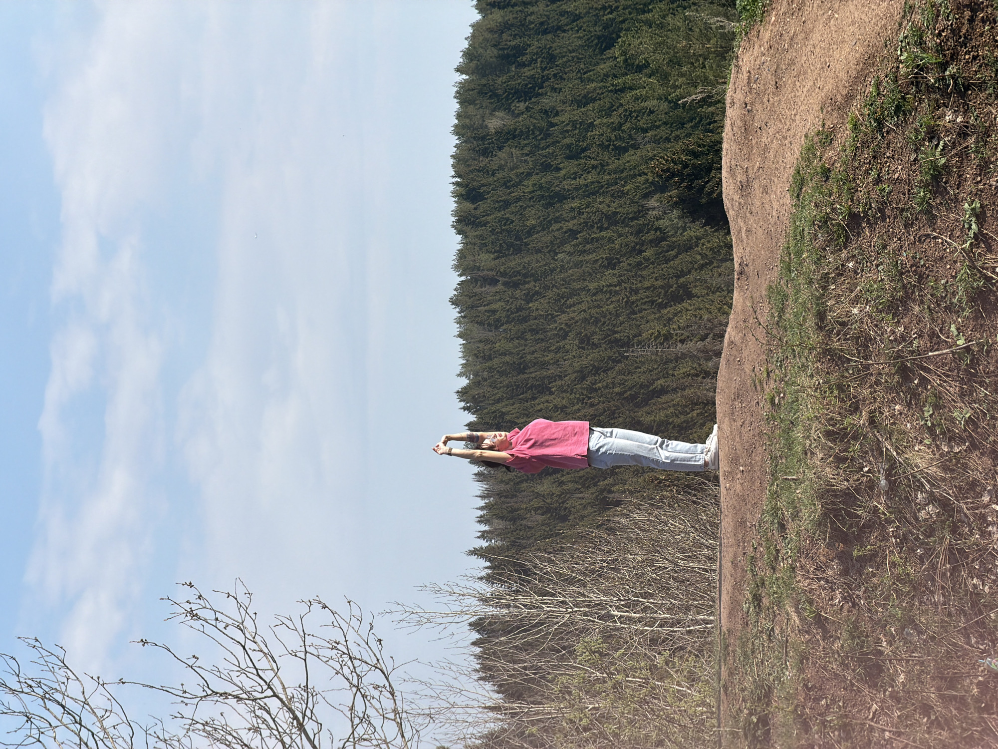 A person in a pink shirt and jeans is standing on a dirt embankment with a dense forest behind them. The sky is blue with scattered clouds. The person is raising their arms above their head. Some bare trees are visible in the foreground. The location appears to be a natural area or park, possibly a wooded hillside, within Perm, Russia.