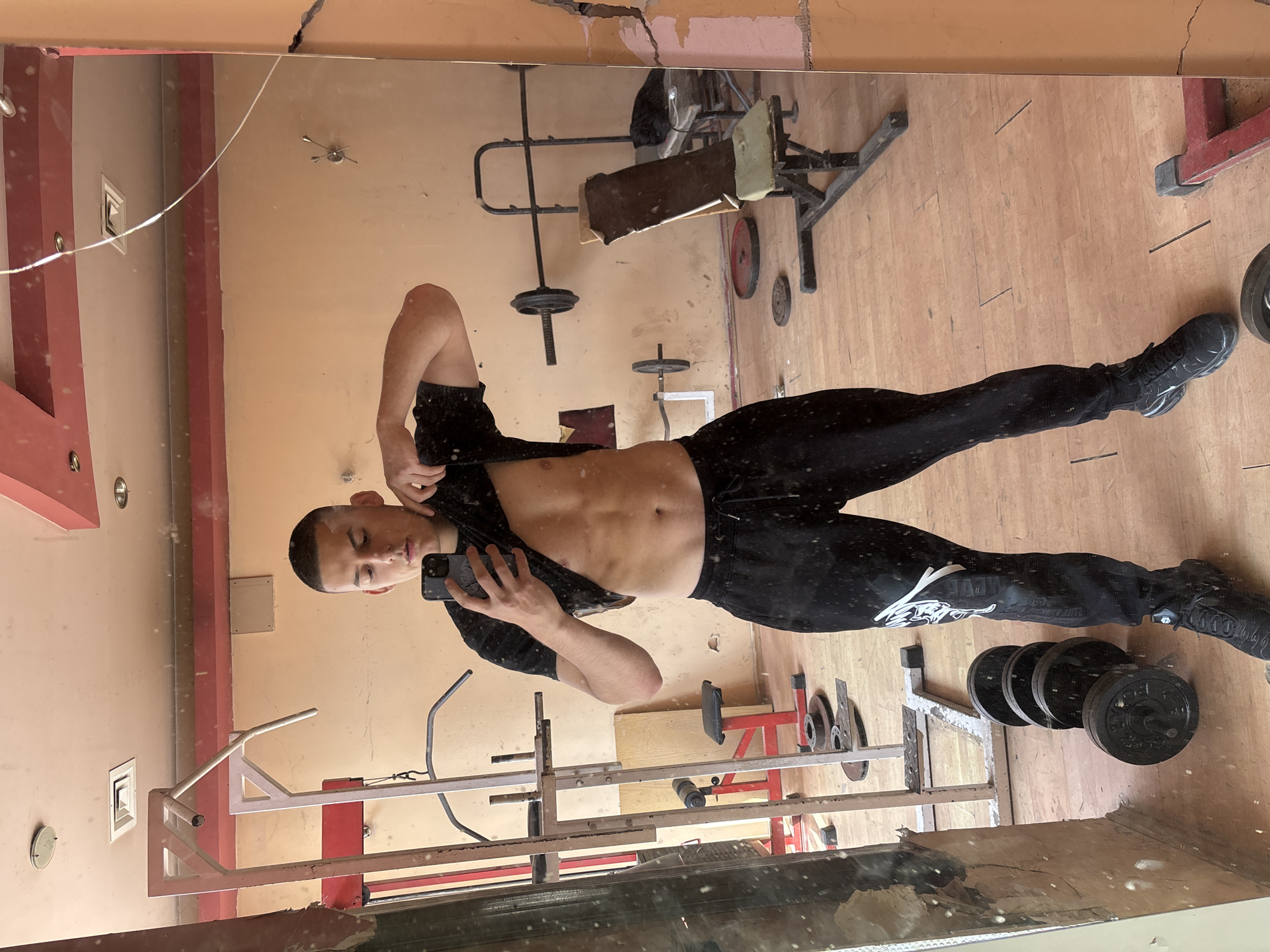 A young man takes a mirror selfie in what appears to be an indoor gym in Goritsa, Bulgaria. He stands facing the mirror, holding a smartphone in his right hand to capture the reflection. He is wearing a black t-shirt, which he has lifted with his left hand to reveal his toned abdomen, and black sweatpants and sneakers.
The gym setting is reflected in the mirror, showing various pieces of workout equipment. In the background, there's a weight bench, what looks like parts of a squat rack or multi-gym frame, and several weight plates on the floor and stacked on a rack. The floor is made of light-colored wood, and the walls are painted a light brown or peach color. The room appears somewhat aged, with visible cracks in the ceiling/upper wall area, and some dirt/smudges on the mirror surface. The equipment also shows signs of wear.
The man's black sweatpants feature a white graphic logo on his left leg (as seen in the reflection), which is stylized and difficult to decipher completely. There's also a small white tag or logo visible on his right shoe.