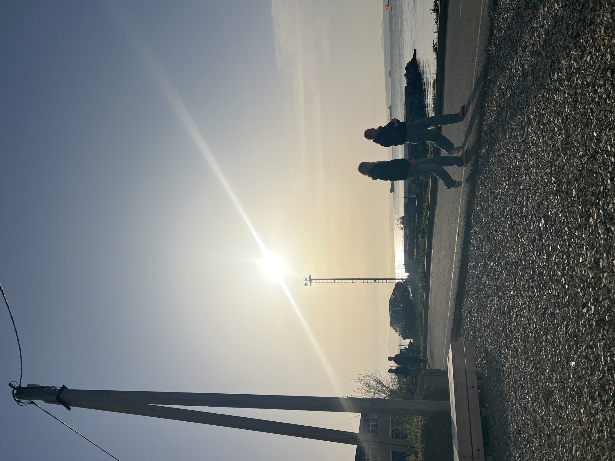 The image depicts a scene in Vladivostok, Russia, characterized by strong backlighting from the sun. In the foreground, a wooden utility pole with attached wires is partially visible against a clear sky. Moving towards the center, the sun is a dominant feature, creating a bright glare. Along a road bordering a body of water, possibly the sea, two individuals are walking. Beyond them, near the water's edge, there are silhouetted structures and a vehicle resembling an excavator. The road's edge transitions into a gravelly area in the lower right corner. The scene conveys a sense of urban or semi-urban environment near the coast.