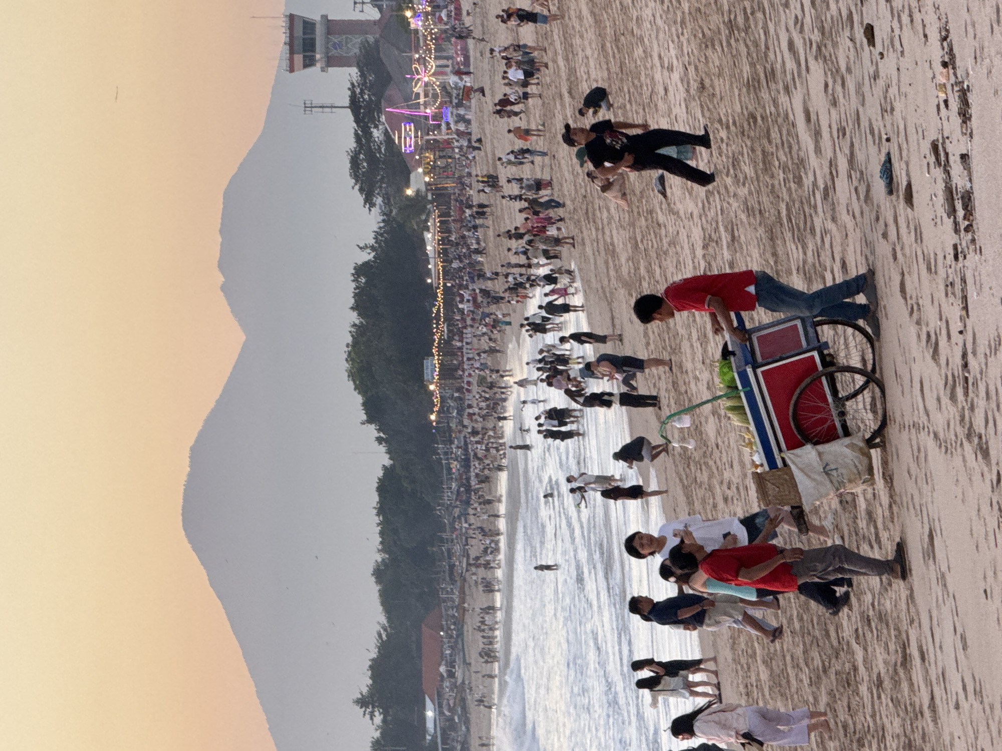 The image depicts a crowded beach scene in Kuta, Indonesia. Numerous people are walking along the beach and near the water's edge. A mountain is visible in the background, along with a structure reminiscent of a lifeguard tower. A vendor with a red cart is seen amongst the beachgoers. The overall lighting suggests it is either early morning or late afternoon.