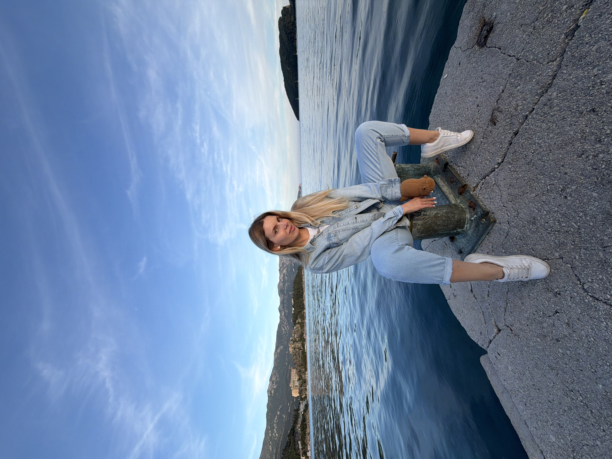 A woman is sitting on a bollard on a concrete pier in Budva, Montenegro. She has long blonde hair and is wearing a denim jacket and jeans, and white sneakers. She is holding a brown stuffed animal. The sea is in the background, with a mountain range visible in the distance. The sky is blue with light clouds.
