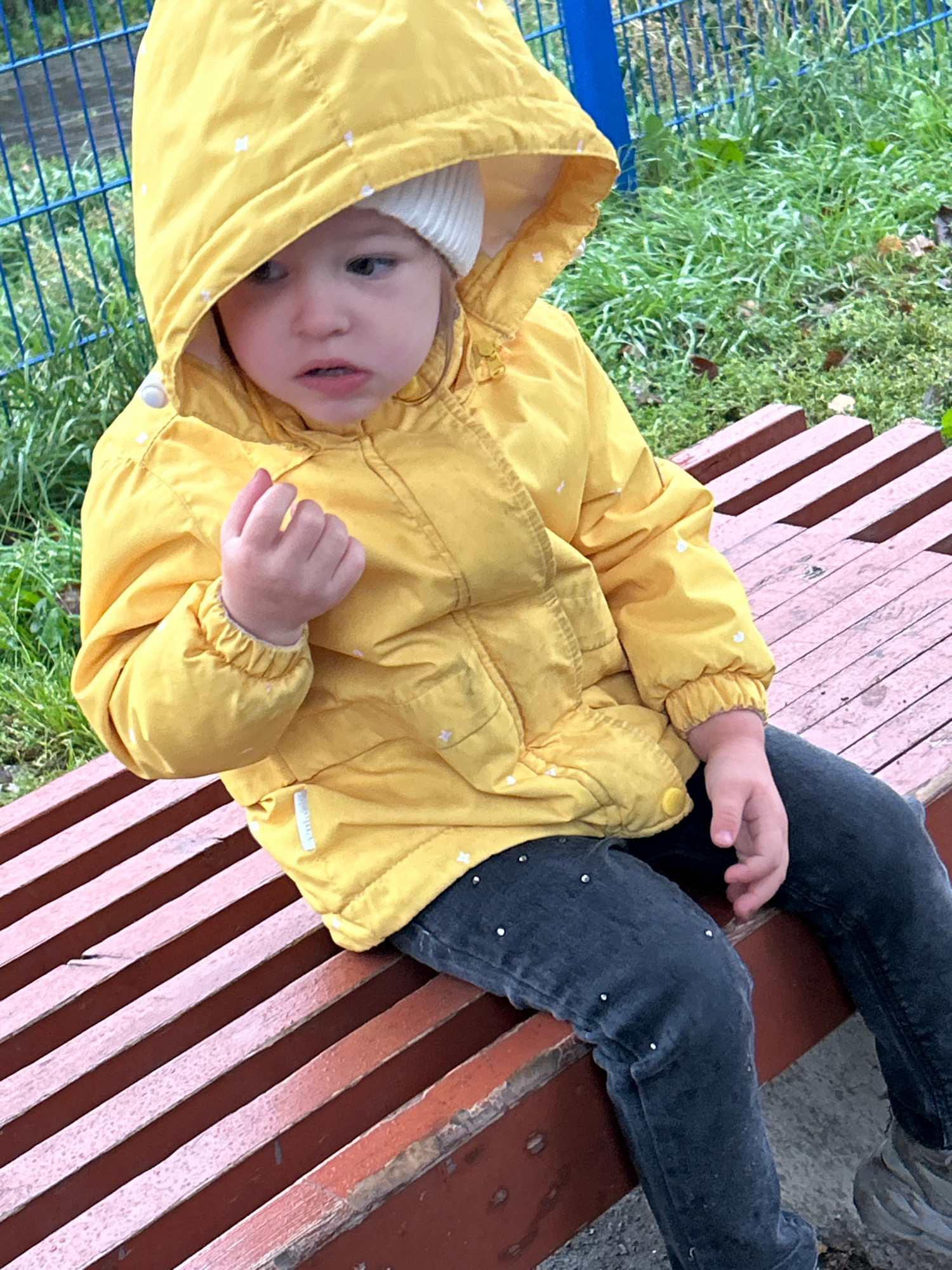 A young girl with fair skin and brown eyes, wearing a yellow hooded jacket with white star patterns and a white knitted hat, sits on a wooden bench. She is wearing dark jeans with small white studs. Her left hand is raised and curled into a fist. The bench is made of wooden slats and is painted a reddish-brown color. In the background, there is green grass and a blue metal fence. The scene appears to be outdoors, possibly in a park or playground. There are no identifiable landmarks that place the image specifically in Tomsk, Russia.