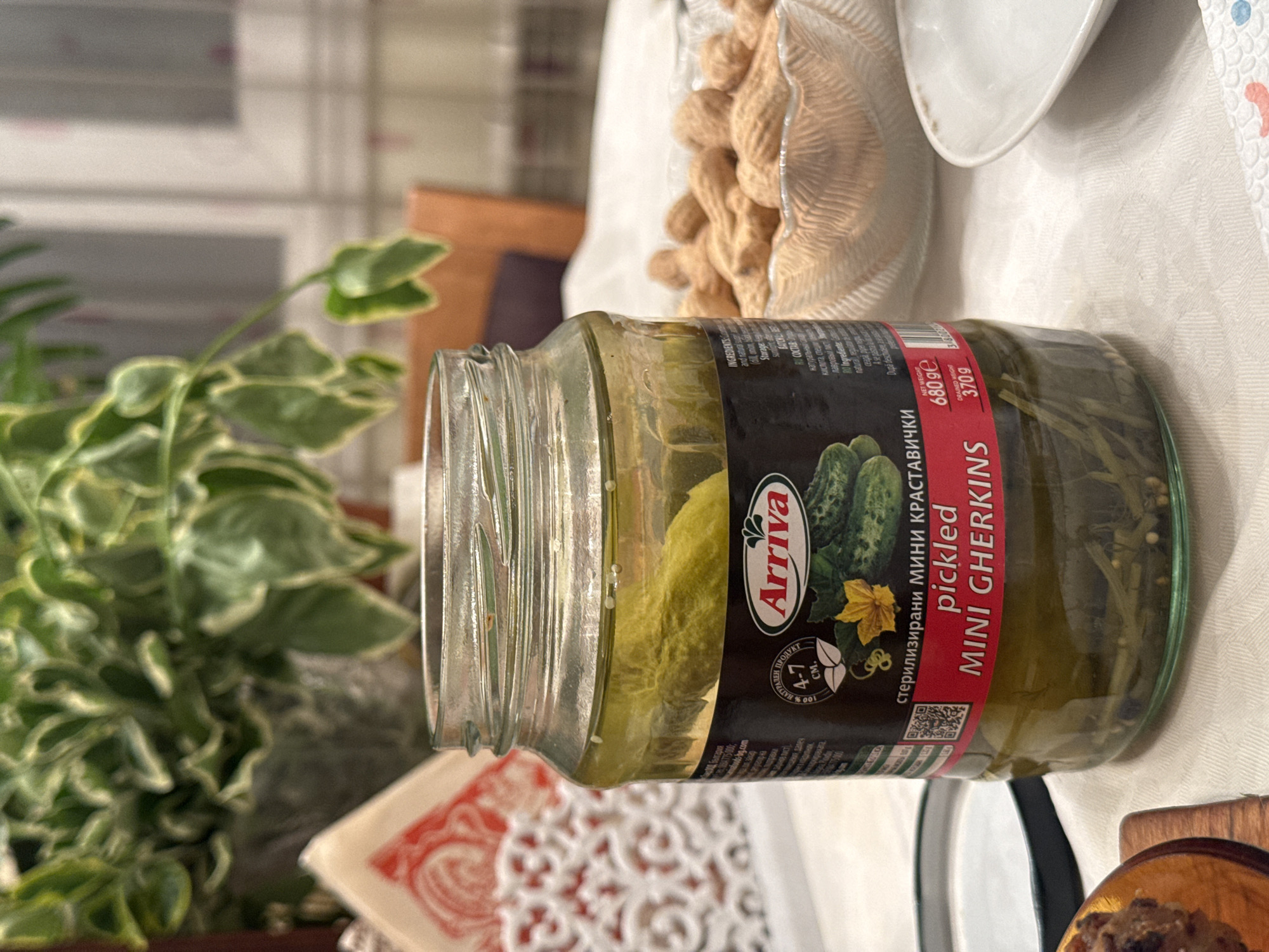 A glass jar of "Arriva" pickled mini gherkins sits on a table. The jar is partially obscured by a plant to the left and a bowl of peanuts and other dishes to the right. The label on the jar includes Bulgarian and English text, stating "стерилизирани МИНИ КРАСТАВИЧКИ" and "pickled MINI GHERKINS". The net weight is marked as 680 ge and the drained weight is 3709. The environment implies a dining or living area. The exact location in Sofia, Bulgaria cannot be determined from the provided image.