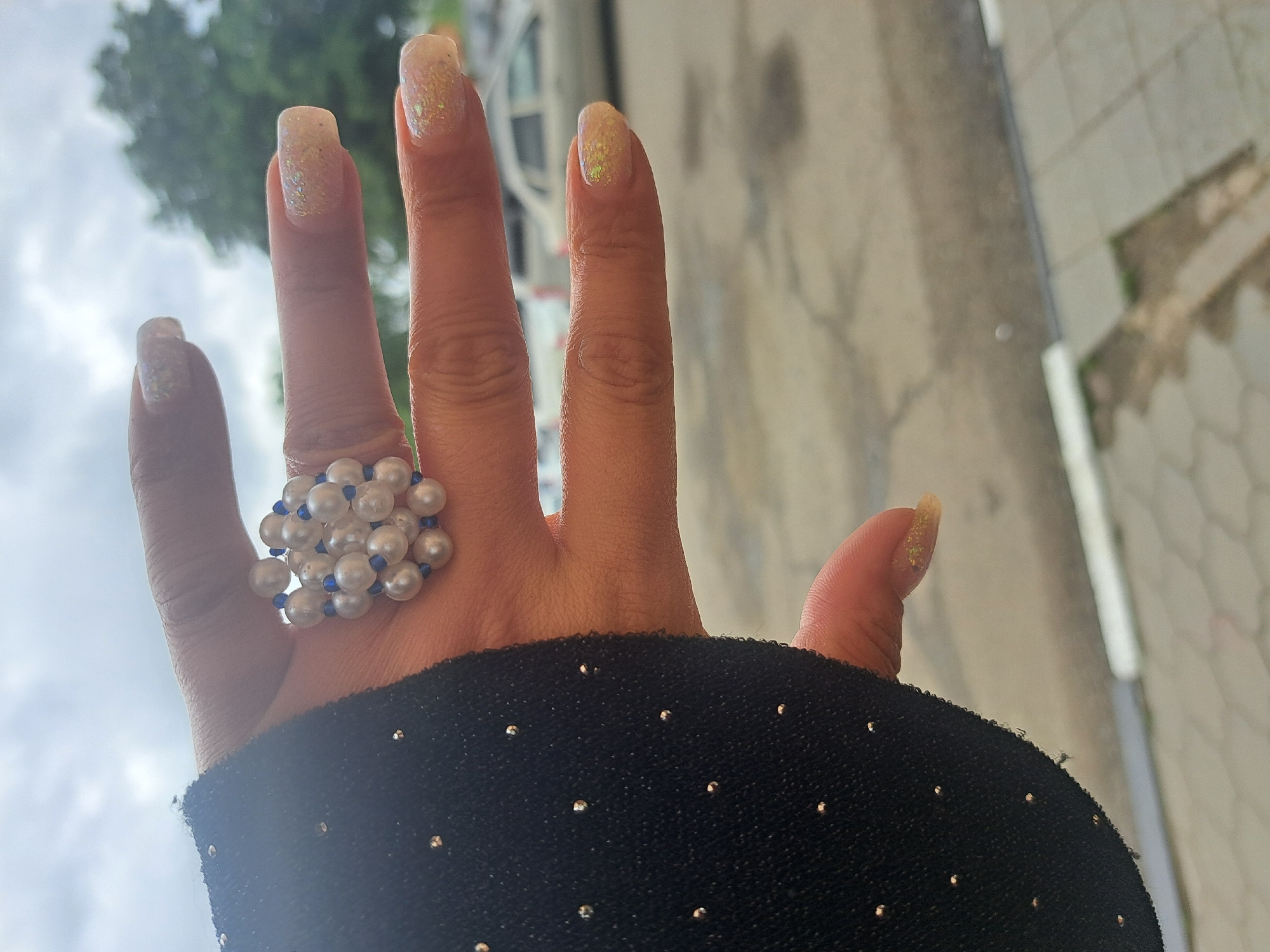 A hand wearing a large ring made of white pearls and small blue beads is presented in the foreground. The hand is adorned with painted nails that have a light base color and a glittery topcoat. The hand is emerging from a black fabric, potentially a sleeve, that is decorated with small, evenly spaced silver studs. In the background, the environment consists of blurred elements: a patch of green possibly representing a tree, a building with white elements, and a textured gray surface, possibly a sidewalk or wall. The overall lighting suggests it is daytime. The location is not identifiable as Leningradskaya, Russia based on the provided image.