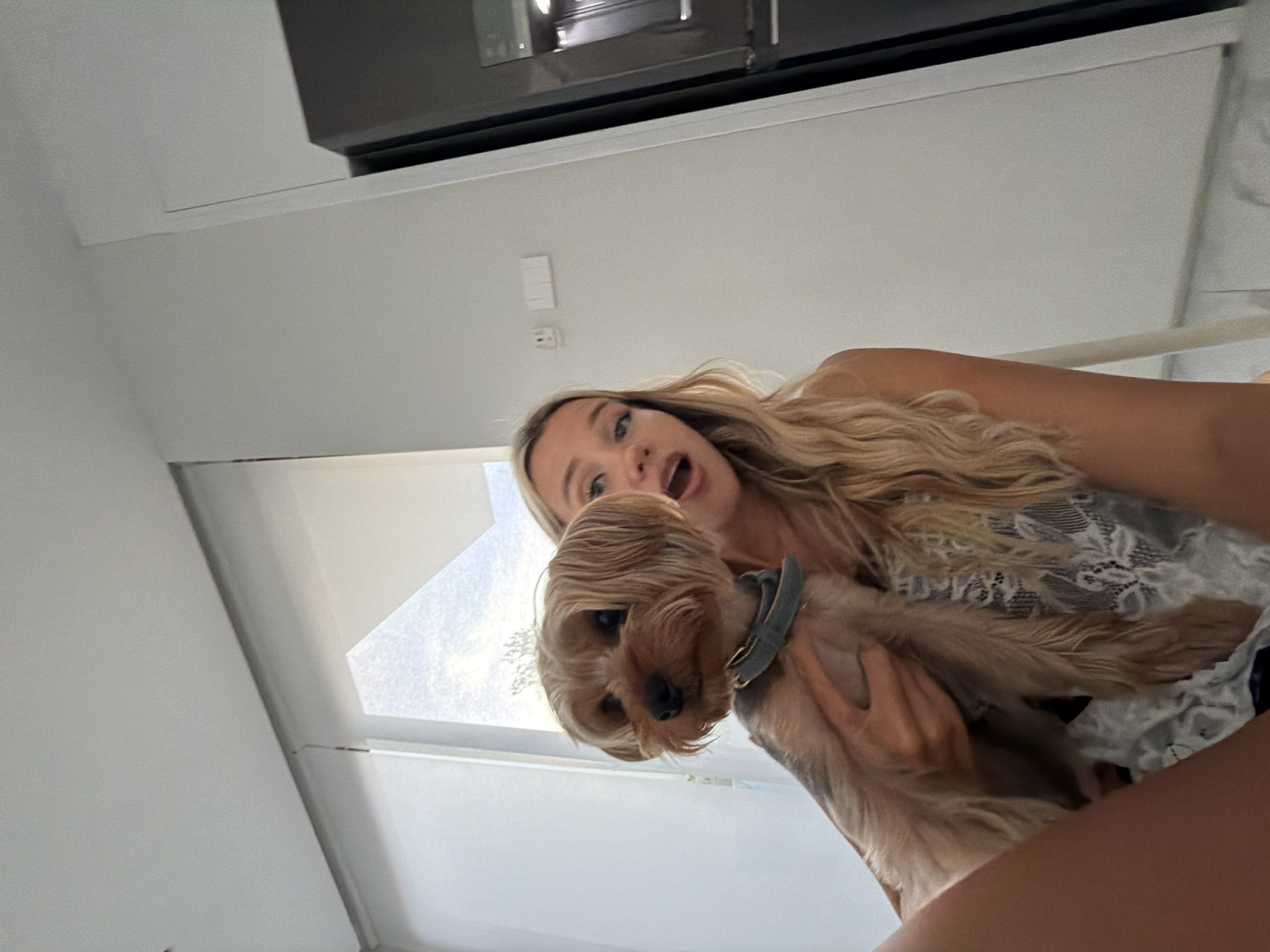 A young woman with blonde hair is holding a small, tan dog with a grey collar. The woman is looking at the camera with her mouth open, and the dog is looking to the left. They are indoors, with a window with sheer blinds in the background. There is also a dark appliance, possibly a refrigerator, visible on the right side of the image. The overall environment appears to be a modern interior with white walls and a minimalist aesthetic. The image has a bright, natural light coming from the window. The location is not specified as Thep Krasatti, Thailand.