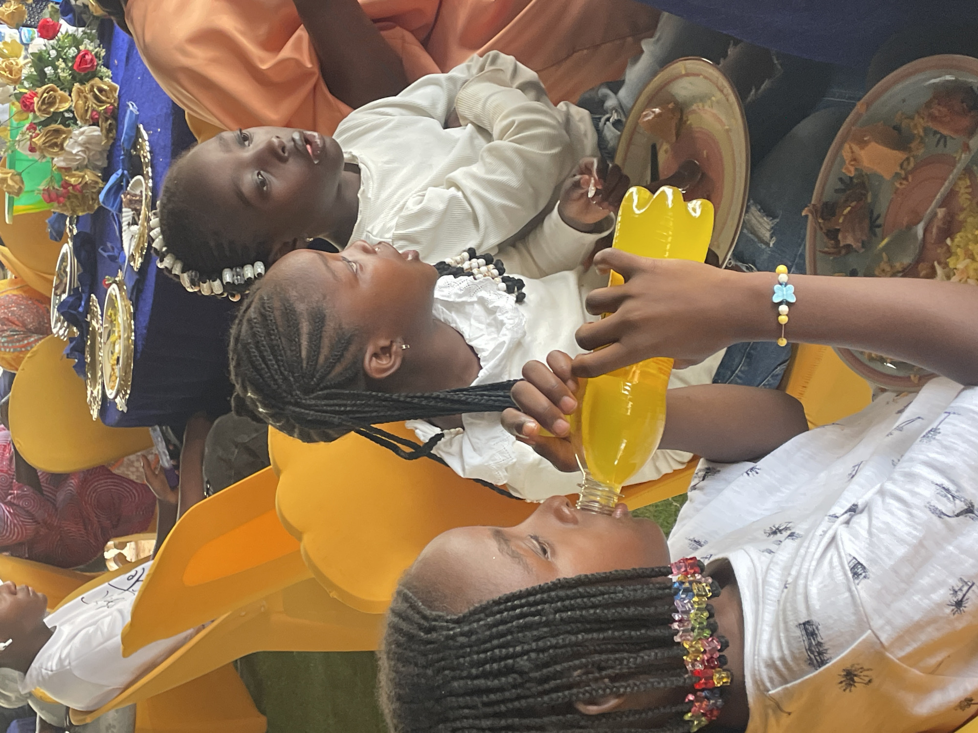 Three girls are seated outdoors, dressed in white clothing and adorned with beaded accessories. The girl at the bottom is drinking from a yellow bottle with assistance. Plates of food are nearby. One of the girls looks upward.