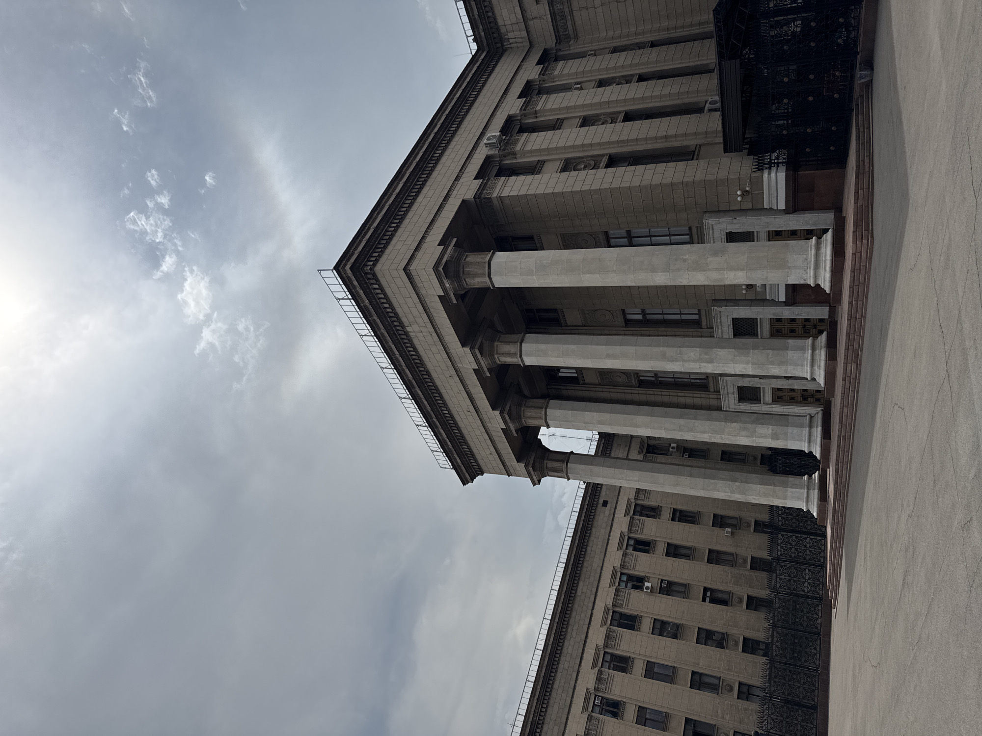 This is a photograph of the Kazakhstan National Academy of Sciences in Almaty, Kazakhstan, taken from a low angle looking upwards. The sky is overcast with thin clouds, and the sun is faintly visible through them. The building itself is a grand, monolithic structure made of light-colored stone. It features a prominent portico with several large, fluted columns supporting a heavy entablature. The facade is characterized by repetitive rows of windows and decorative elements. To the left, another section of the building is visible, with a similar architectural style. A dark, ornate metal fence runs along the front of the property. The ground in front of the building is paved with concrete or asphalt. The overall impression is one of imposing architecture and a somewhat somber atmosphere due to the cloudy sky.