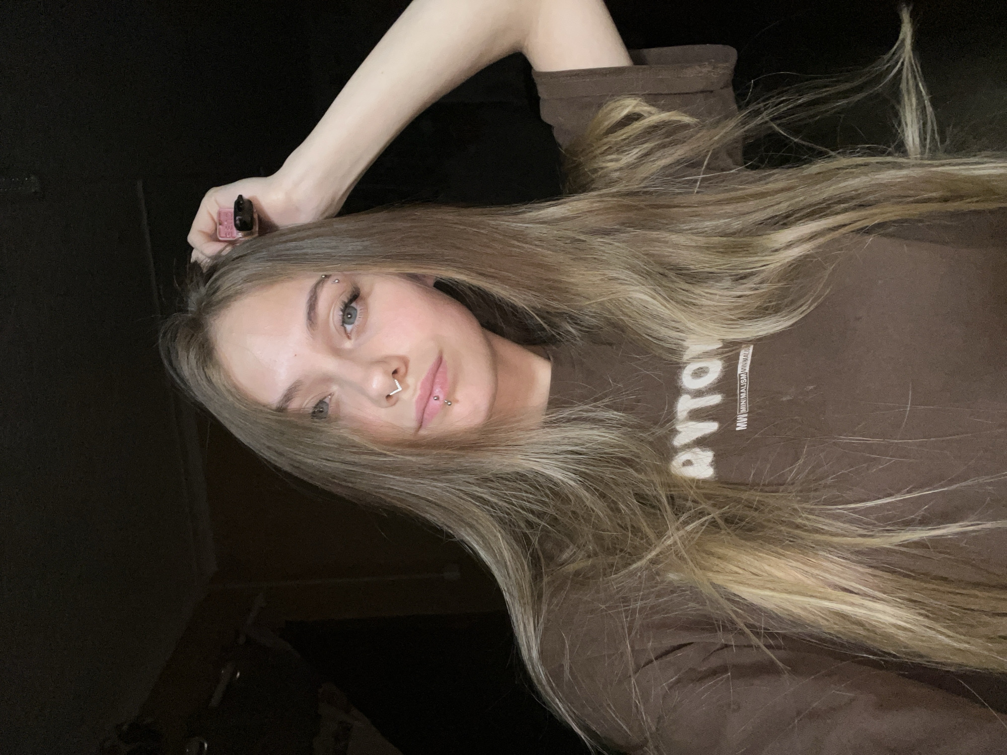 A young woman with light brown hair, eyebrow piercings, a nose ring, and a lip piercing is posing for a selfie. She is wearing a brown shirt with white lettering. Her right arm is raised above her head, holding a small, pink device. The background is dark. The location is not discernible from the image.