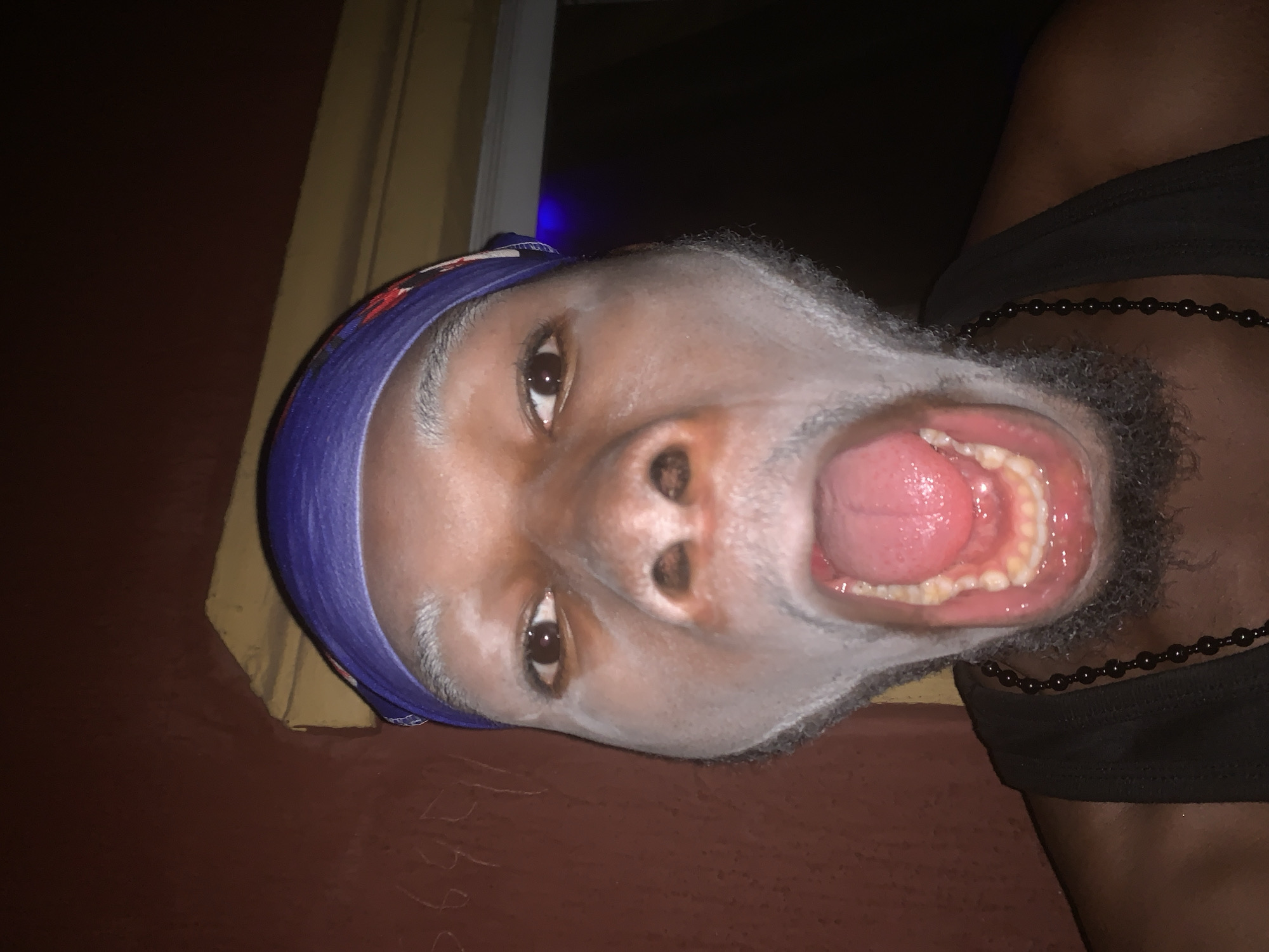 An upward-angled, close-up shot of a person, likely male, with dark skin, is captured in a dimly lit indoor setting, possibly at night, in Onitsha, Nigeria. The individual is wearing a blue patterned head covering and a dark, possibly sleeveless, top, adorned with a black beaded necklace. They have visible stubble or a short beard on their face. The person looks directly into the camera with wide, open eyes and their mouth agape, revealing their tongue and teeth, which could suggest an expression of surprise, a yell, or a playful pose. The background on the left features a dark brown wall and a lighter yellow or beige vertical element, possibly a curtain or window frame. On the bottom right, a reddish-brown surface, possibly a wall or furniture, shows faint, unidentifiable markings.