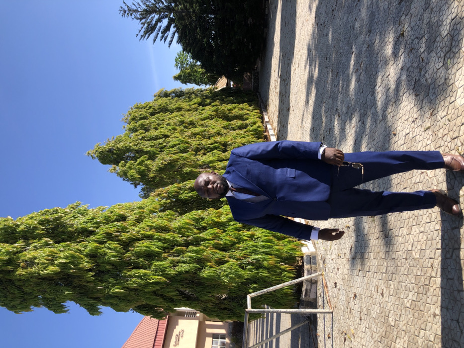 A man in a navy blue suit and brown shoes stands on a stone path next to a large, vibrant green bush. He holds eyeglasses in his hand. A building with a red roof is visible in the background to the lower left. The sky is clear and bright blue. Trees are visible in the background.