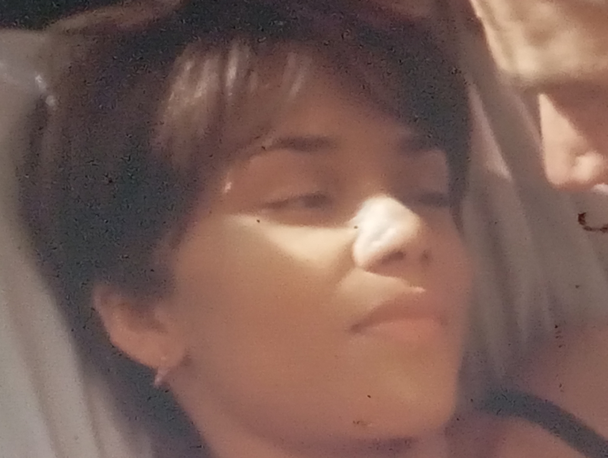 The image is a close-up, grainy, and slightly out-of-focus photograph of a person lying down. The person has short, dark hair and their face is illuminated from the side, creating shadows. Their eyes are closed or looking away from the camera, and their mouth is slightly open. A small earring is visible on their left ear. The background is out of focus, but appears to be a light-colored surface, possibly bedding or fabric. There are some dark specks visible throughout the image, which could be dust or artifacts. No specific location in Sofiivska Borshchahivka, Ukraine, can be identified from the image.
