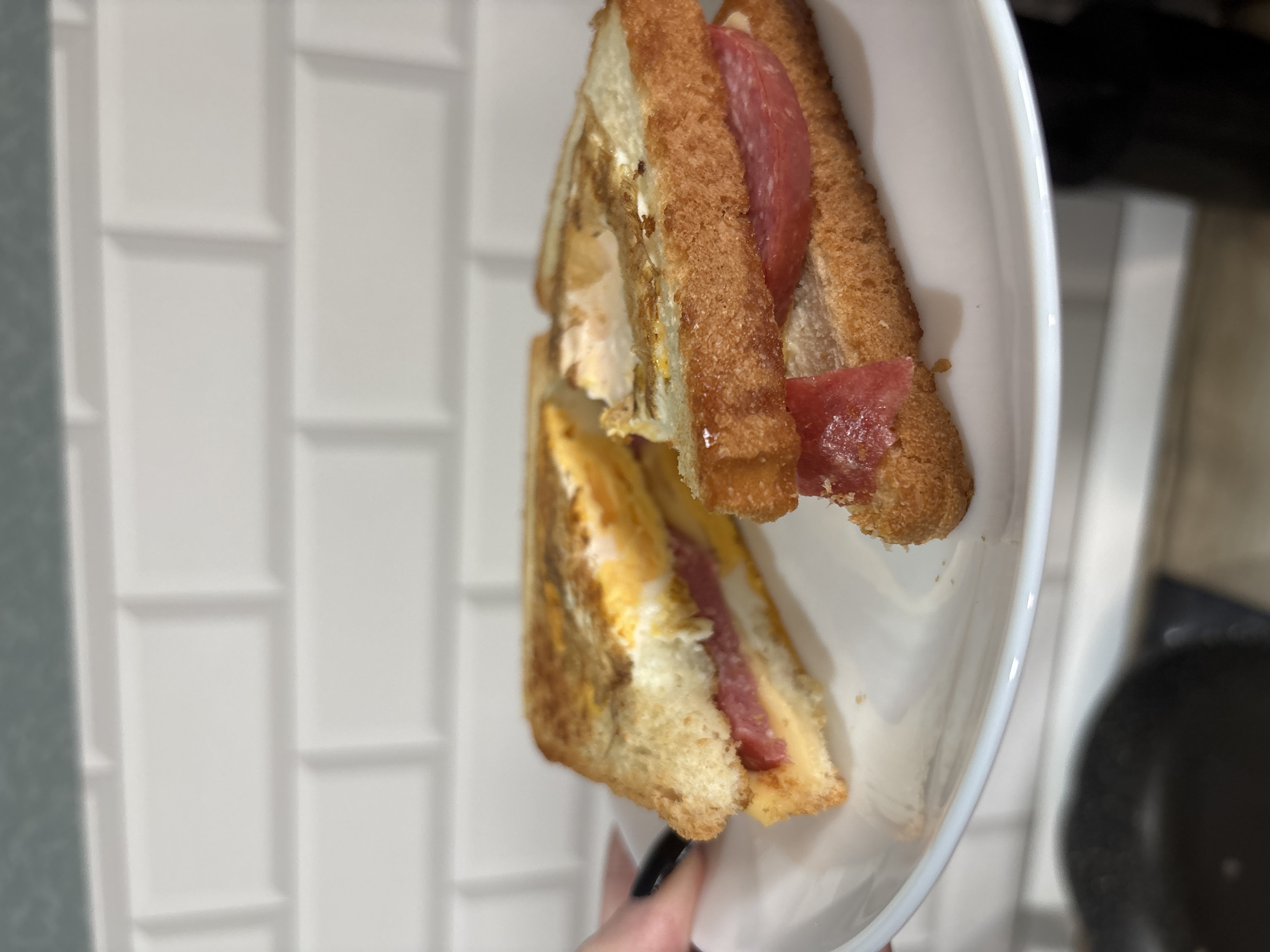 The image shows a sliced sandwich on a white plate. The sandwich appears to contain egg and salami. The background includes white subway tiles and a blurred view of what looks like a kitchen counter with a stovetop and a pan. The presence of the subway tiles and the kitchen setting suggest the photo was taken indoors. There is no information in the image that can confirm its location as Saint Petersburg, Russia, or provide further details about people or interactions.