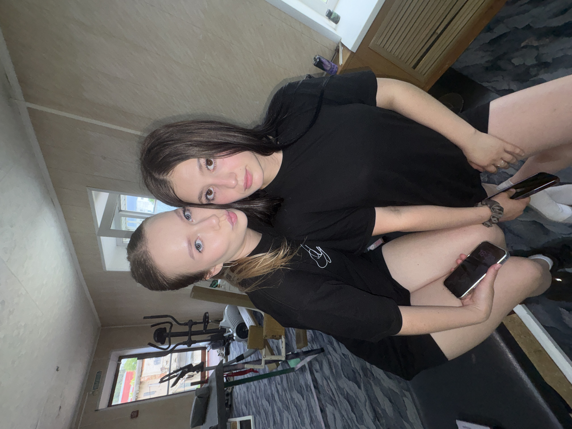Two young women are posing for a photo in what appears to be a home gym or exercise room. The room has exercise equipment, including an elliptical machine and a weight machine, visible in the background. The floor is covered in a patterned rug. The walls are paneled with light-colored wood. There is a window with a view of buildings and greenery. The woman on the left is wearing a black t-shirt and shorts, and she is holding a smartphone in her hand. The woman on the right is also wearing a black t-shirt and shorts. They are both looking at the camera. The image was taken in Chaikovsky, Russia.