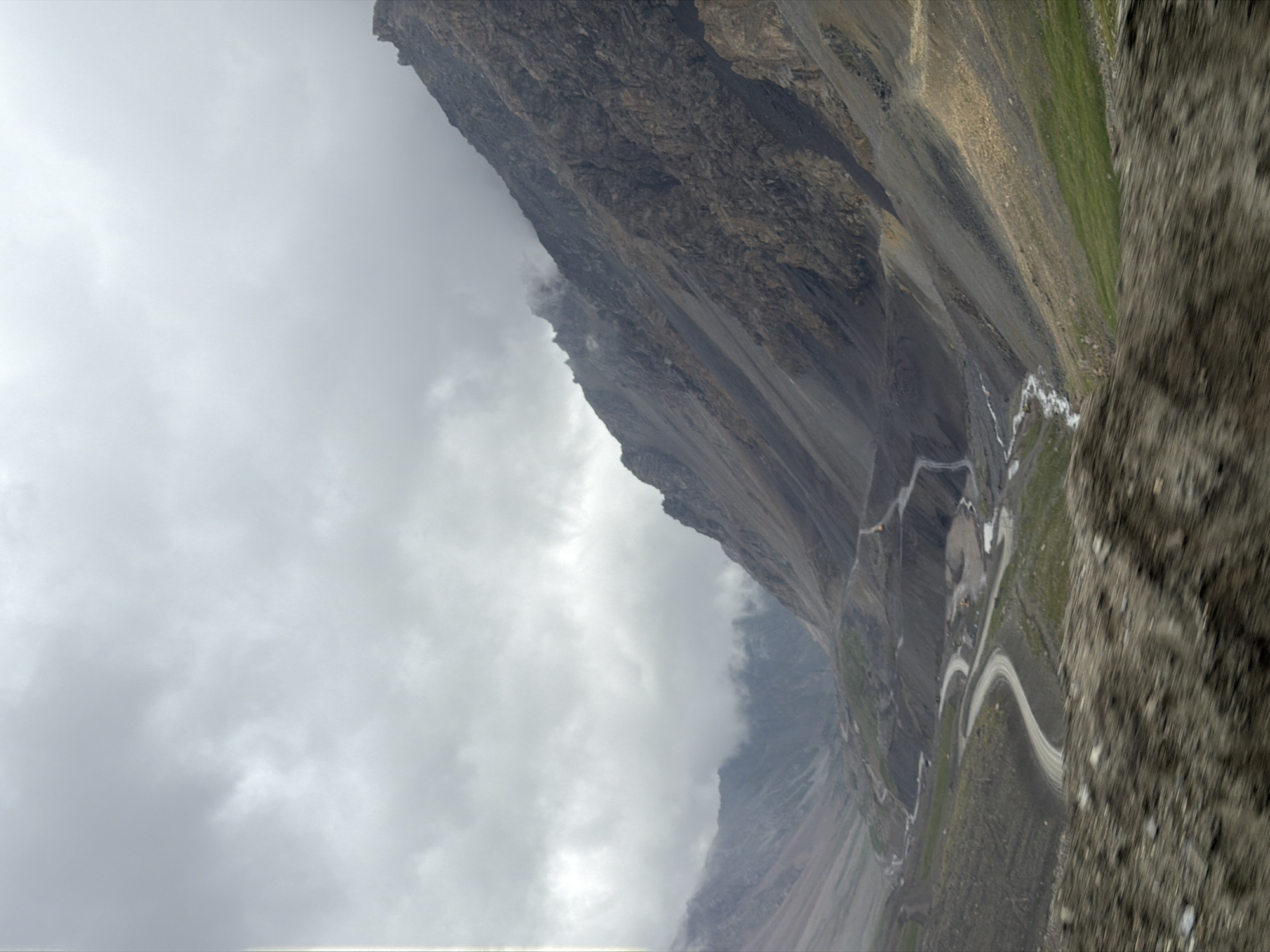 The image shows a desolate mountain landscape in Kyrgyzstan under a cloudy sky. A winding dirt road snakes through a valley, flanked by steep, rocky slopes. Some patches of green grass are visible on the lower slopes, contrasting with the barren, grey terrain. The road appears to be under construction or maintenance, with some yellow construction equipment visible in the distance. The overall impression is one of remoteness and rugged natural beauty.