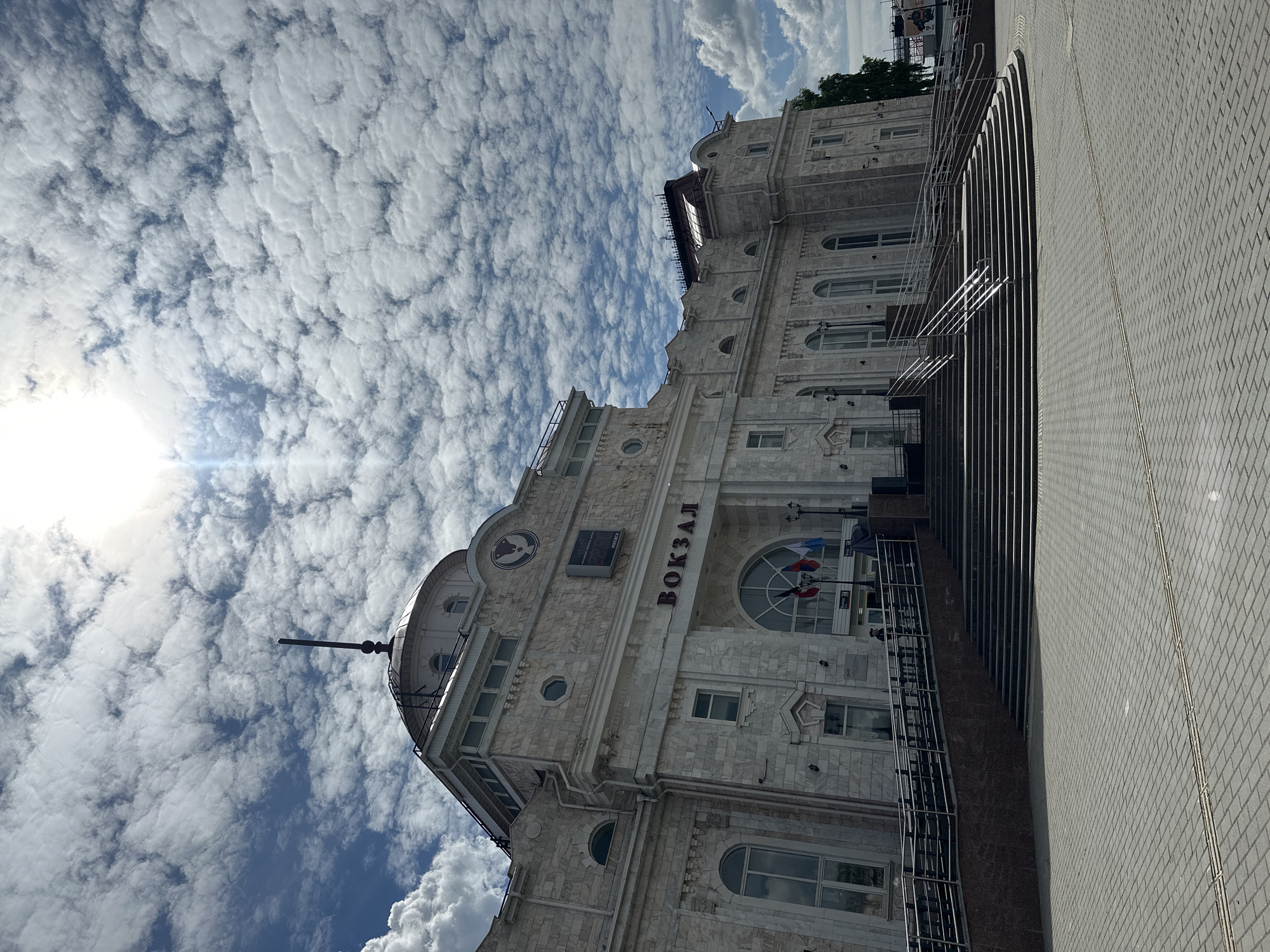 This image depicts the main facade of the Izhevsk train station (Vokzal) in Izhevsk, Russia, under a bright, partly cloudy sky. The building is constructed of light-colored, possibly marble or granite, stone cladding, featuring classical architectural elements such as pilasters, arched windows, and decorative cornices.
Central to the facade, above the main entrance, is the word "ВОКЗАЛ" (Vokzal) in prominent red capital letters. Above this, a digital clock displays "22:00" and "ИЖЕВСК" (Izhevsk). To the left of the clock, a circular emblem featuring a bird-like figure is visible. The main entrance area includes a large arched window, and directly below it, three flags are displayed: the flag of Russia, and two other regional flags (likely Udmurtia and Izhevsk). A tall, dark spire or finial tops a dome on the left side of the building's roofline.
In the foreground, a large plaza paved with grey interlocking pavers extends towards the viewer. A wide, multi-tiered set of stairs with metal railings leads up to the station's main entrance. To the left of the stairs, a gently sloping ramp with similar metal railings provides accessible entry. The base of the stairs and the building's foundation are clad in a dark, reddish-brown stone. Golden tactile paving strips are visible on the grey plaza in the lower center.
One individual in dark clothing is observed ascending the accessibility ramp on the left side of the building's entrance area. The sun, positioned in the upper center-right, casts strong light, creating a lens flare and distinct shadows on the ground and parts of the building, indicating a sunny day. The sky is largely covered with numerous small, lumpy cumulus clouds scattered across a blue background. A tree with green foliage is partially visible on the far right side of the frame.