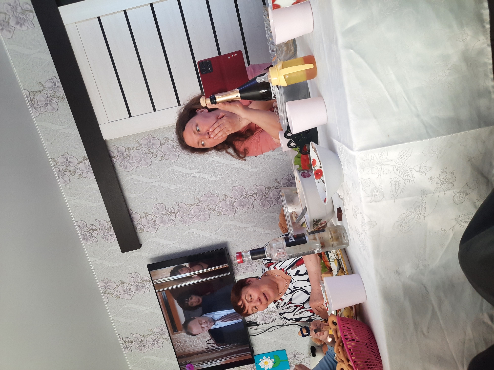 The image shows an indoor scene with a wall featuring floral wallpaper and a light-colored door with dark trim. A television is visible on the wall, displaying an image of several people. In front of the television, an elderly woman is sitting at a table covered with a white tablecloth. On the table, there are various items including pink cups, a bottle of vodka, a pink basket, and other objects.
Across from the woman, another person is visible holding a champagne bottle and a baby bottle. A red phone case is visible nearby. The overall scene suggests a gathering or celebration. It is not possible to determine the location in Krasnogorskoye, Russia from this image alone.