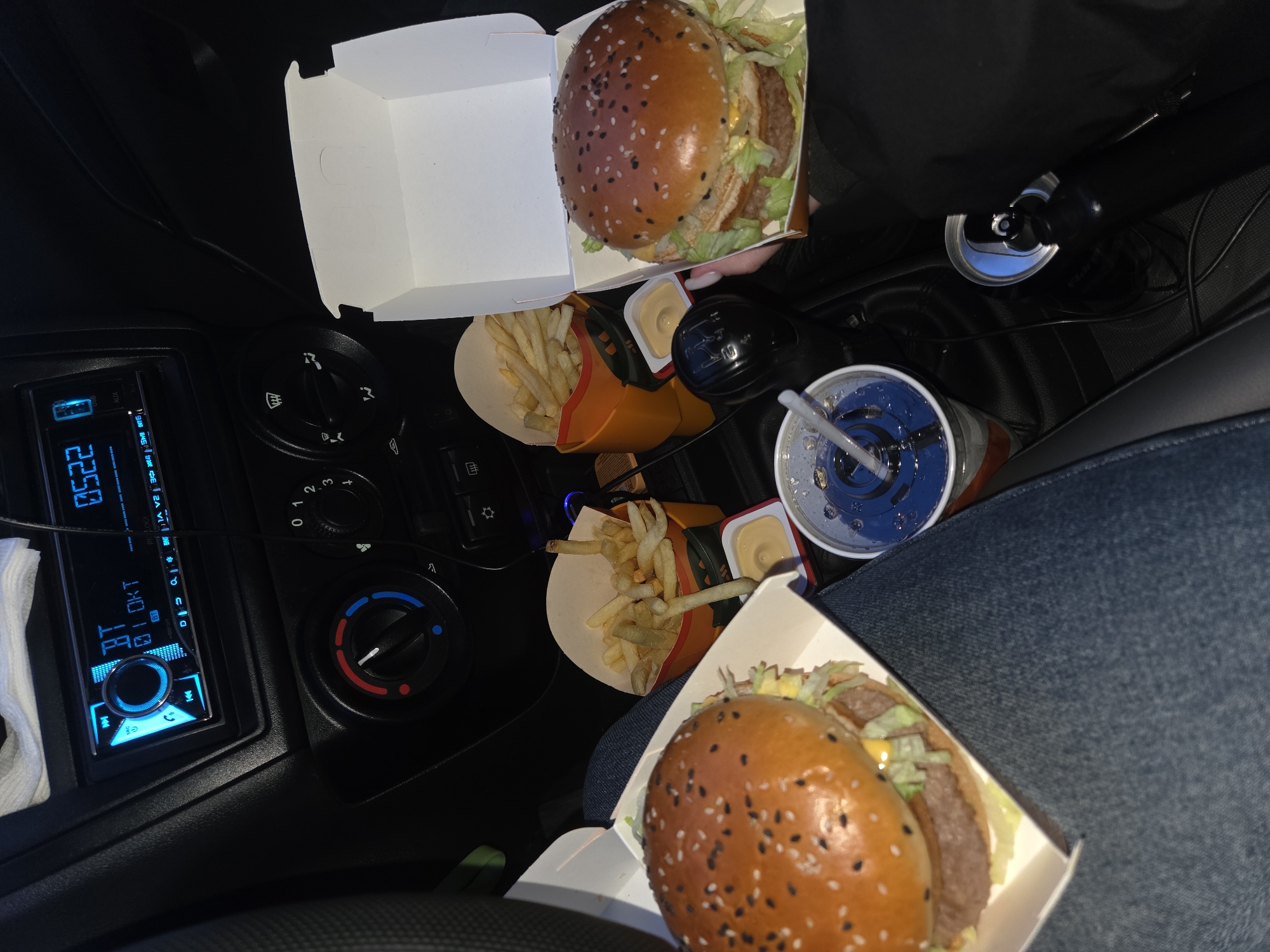 The image shows a cluttered car interior filled with McDonald's fast food. Two Big Macs sit in open white cardboard boxes, with sesame seed buns and visible lettuce and meat patties. Two cartons of McDonald's fries, partially eaten, are present with small containers of dipping sauce. A cup of soda with a blue lid and straw is visible, along with a can of soda. The scene is likely inside a vehicle, as indicated by the car's dashboard and gear stick. The dashboard features an aftermarket car radio, displaying the numbers "0522" and the letters "BT OKT,". Parts of jeans are visible, presumably belonging to a person in the car. The exact location in Perm, Russia cannot be ascertained from the provided image.
