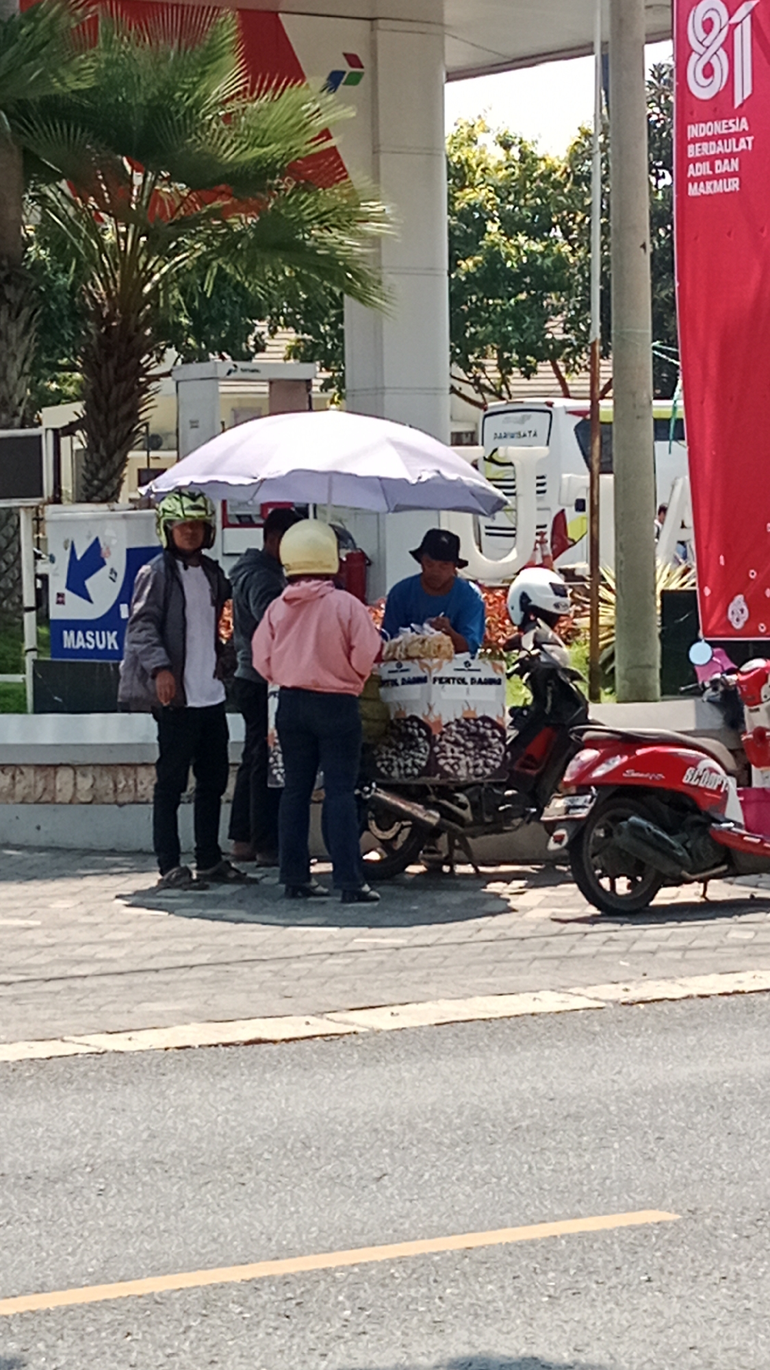 At a roadside in Banyuglugur, Indonesia, a vendor operates a small cart selling snacks from under a large white umbrella. A man in a blue shirt and a black hat stands behind the cart, attending to customers. Two individuals on the left, wearing helmets and casual attire, are interacting with the vendor. A red motorcycle with a "SCOOPY" label is parked next to the cart, along with another dark-colored motorcycle. In the background, a white bus and a sign with the word "MASUK" (enter) are visible, along with palm trees and a tall pole with red signage indicating "81 INDONESIA BERDAULAT ADIL DAN MAKMUR" (81 Indonesia Sovereign Just and Prosperous). The scene takes place on a paved surface next to a road with a yellow line marking.