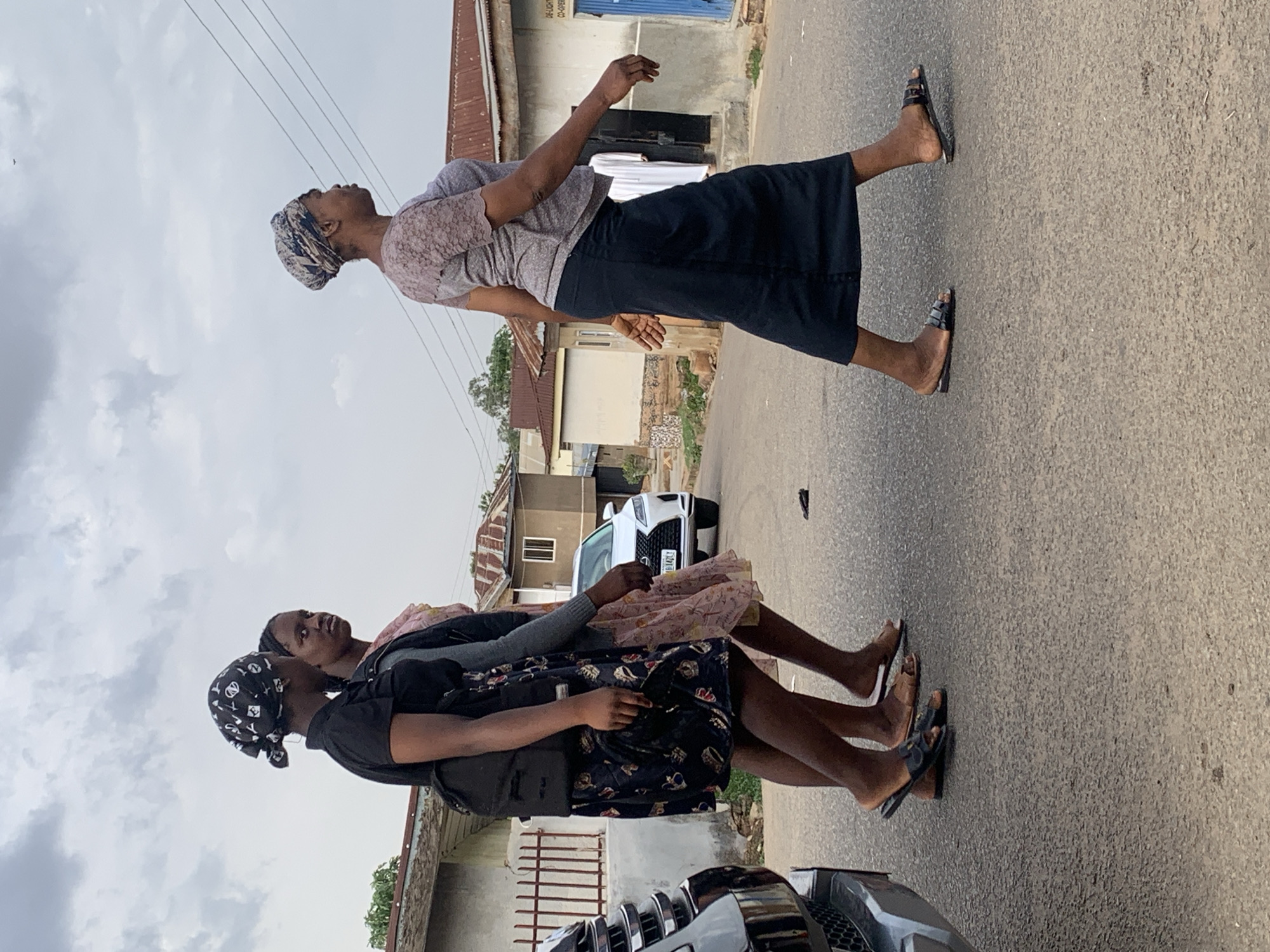 Three African women stand on a paved road in Jos, Nigeria. The woman closest to the camera wears a black patterned dress, a headscarf, a black bag, and black sandals. Another woman, standing slightly behind, wears a light pink dress and a grey top. The third woman, set further away, is attired in a gray top, a navy blue skirt, a headscarf, and black sandals. All three women are looking up toward the sky. Behind them, there is a white car and buildings with corrugated metal roofs. The road is cracked, and overhead power lines are visible. The sky is overcast.