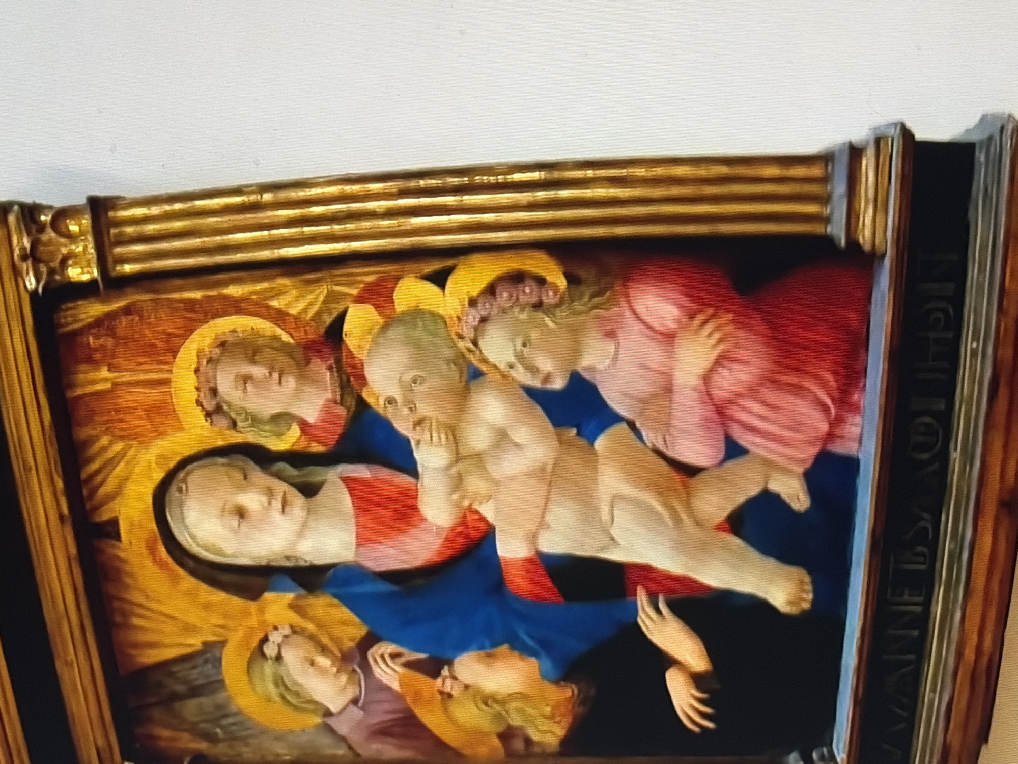 In a painting, the Virgin Mary, depicted with a dark veil over her head, cradles the infant Jesus. She wears a red garment beneath a blue mantle. The infant Jesus, with a pale complexion, is being held and appears to be reaching towards the Virgin Mary's face. To the left of Mary, another child with blond hair, wearing a purple tunic and a floral headband, looks down with hands clasped. Behind Mary, a child with blond hair and a floral headband, wearing a red garment, is partially visible. To the right of the infant Jesus, a child with blond hair, wearing a pink dress and a floral headband, looks upwards. The figures are set against a background of golden rays, suggestive of a divine light. The painting is enclosed in a gilded frame with fluted columns on the right side. The bottom of the frame has an inscription. The painting is displayed against a white wall.