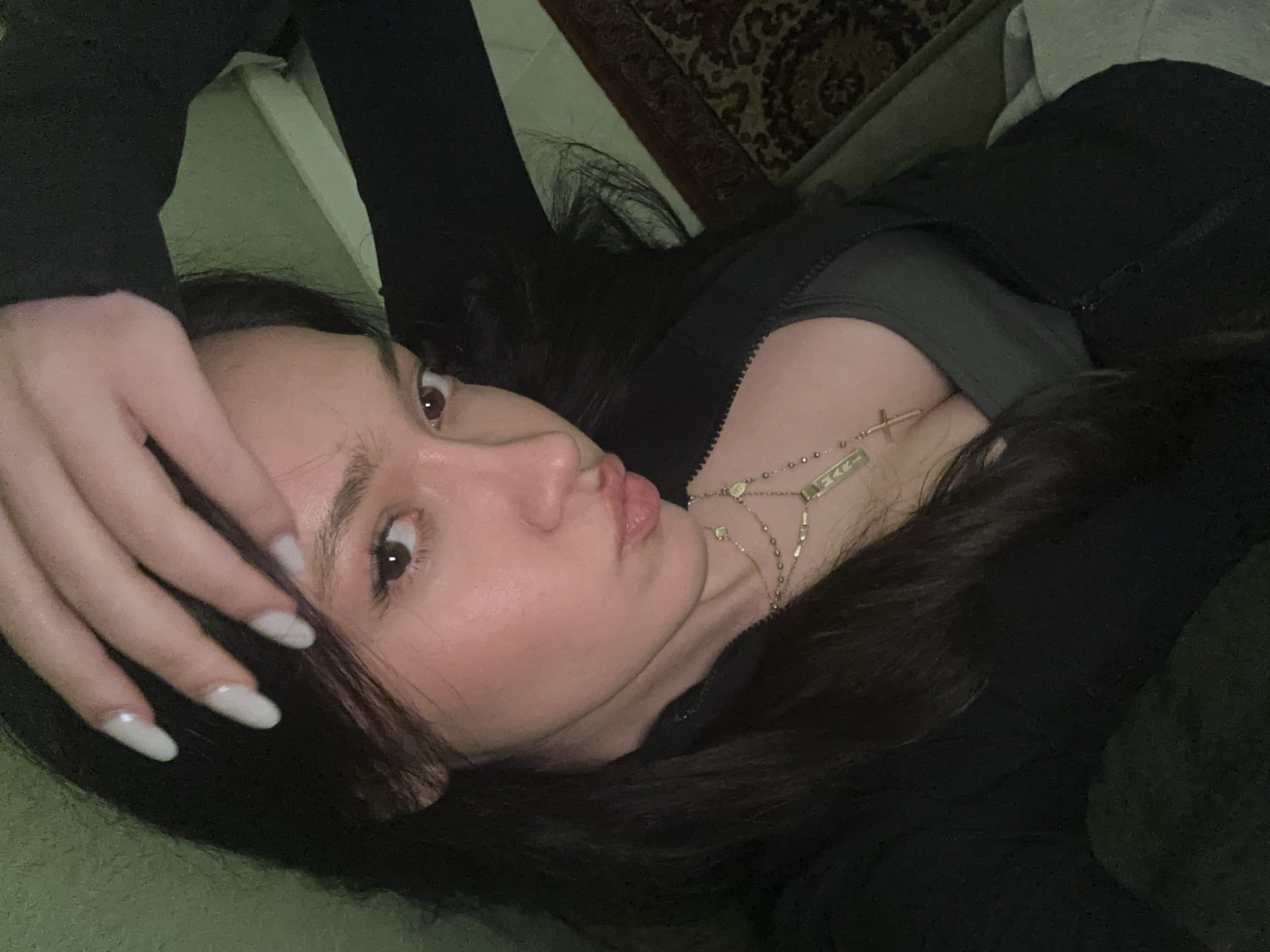 A young woman is captured in a close-up, selfie-style shot, lying down indoors. Her head is tilted, with her face angled towards the viewer, and her dark brown eyes are open and looking directly at the camera. She has long, dark brown hair, styled with a side part, which cascades around her face and over her shoulders. Her lips are slightly pursed in what appears to be a "kiss face" or "duck face" expression. Her skin looks smooth with a natural sheen.
Her left hand, positioned near the left side of her face, has its fingers gently resting on her forehead and hair, partially obscuring her temple. She has long, oval-shaped nails painted a light, opaque color, possibly off-white or very pale grey.
She is wearing a dark, possibly black or very dark grey, zipped top, which is partially unzipped to reveal a glimpse of a darker undergarment, likely a bra or sports top. Adorning her neck and chest are multiple layered gold-colored necklaces. One chain features small beads and a small crucifix pendant. Another chain has a rectangular pendant with text that appears to read "NAK" or "MAK". There are also other delicate chains and small charms visible.
The background suggests an indoor setting. To the upper right, a section of a patterned rug or carpet with dark and lighter brown tones is visible. The rest of the background primarily consists of a solid, muted green color, likely a sofa, bed, or cushion she is resting on. The lighting appears somewhat dim, contributing to a casual and intimate atmosphere.