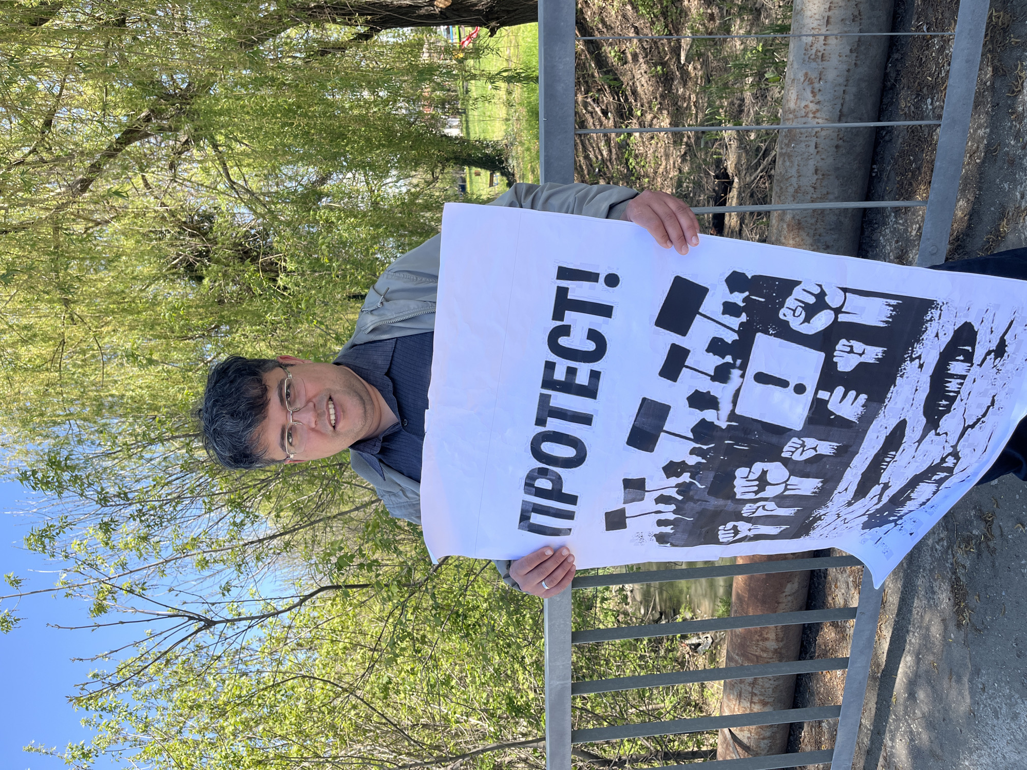 A man with dark hair and glasses is holding a protest sign in Lozarevo, Bulgaria. The sign has the word "ПРОТЕСТ!" in large Cyrillic letters, along with an image of fists raised in solidarity and various signs. The man is standing next to a metal railing, and trees and a body of water are visible in the background. The location is identified as Lozarevo, Bulgaria.