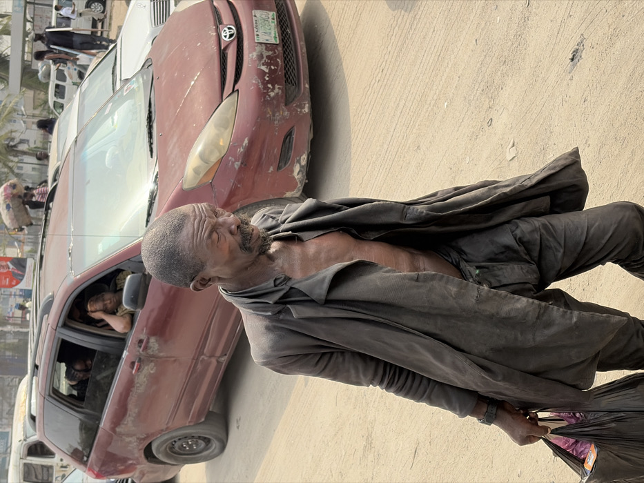 The image depicts a daytime scene on a dusty street in Jakande, Nigeria, with a male subject in the foreground and a line of vehicles in the background.
The primary subject is an older, dark-skinned male, approximately 50-70 years old, with a short, graying beard and mustache, and visible wrinkles on his face. His eyes appear closed or heavily lidded, and he is looking downwards and to his right. He wears a dark, unbuttoned, long-sleeved overcoat or jacket over a bare torso, revealing his chest and abdomen. His dark trousers are also visible, appearing loose and somewhat disheveled. He has a dark-colored wristband on his left wrist and carries two dark plastic bags, one partially open, containing items, in his left hand. The man is walking from left to right across the frame.
Behind the man, a maroon Toyota Sienna minivan (likely a second-generation model, 2003-2010) is prominently featured. The vehicle displays significant paint damage, scratches, and dirt on its front bumper and sides. The license plate, visible on the front, reads "LAGOS LA-239 GD". A male passenger with a light-colored shirt is visible in the driver's side front seat. A female passenger, with reddish-brown hair and a light-colored shirt, is visible through the open front passenger window, resting her arm on the window frame. Other passengers are visible in the rear of the minivan.
Further in the background, to the right of the minivan, a white sedan is partially visible. Additional vehicles and pedestrians are discernible in the distance. One person is seen carrying a large, bundled item on their head. A billboard with a male political figure (likely Bola Ahmed Tinubu, based on similar campaigns) and text is visible in the far background. The street surface is a mixture of worn asphalt and dirt, suggesting an unpaved or poorly maintained road. The lighting indicates bright daylight, possibly harsh sunlight.