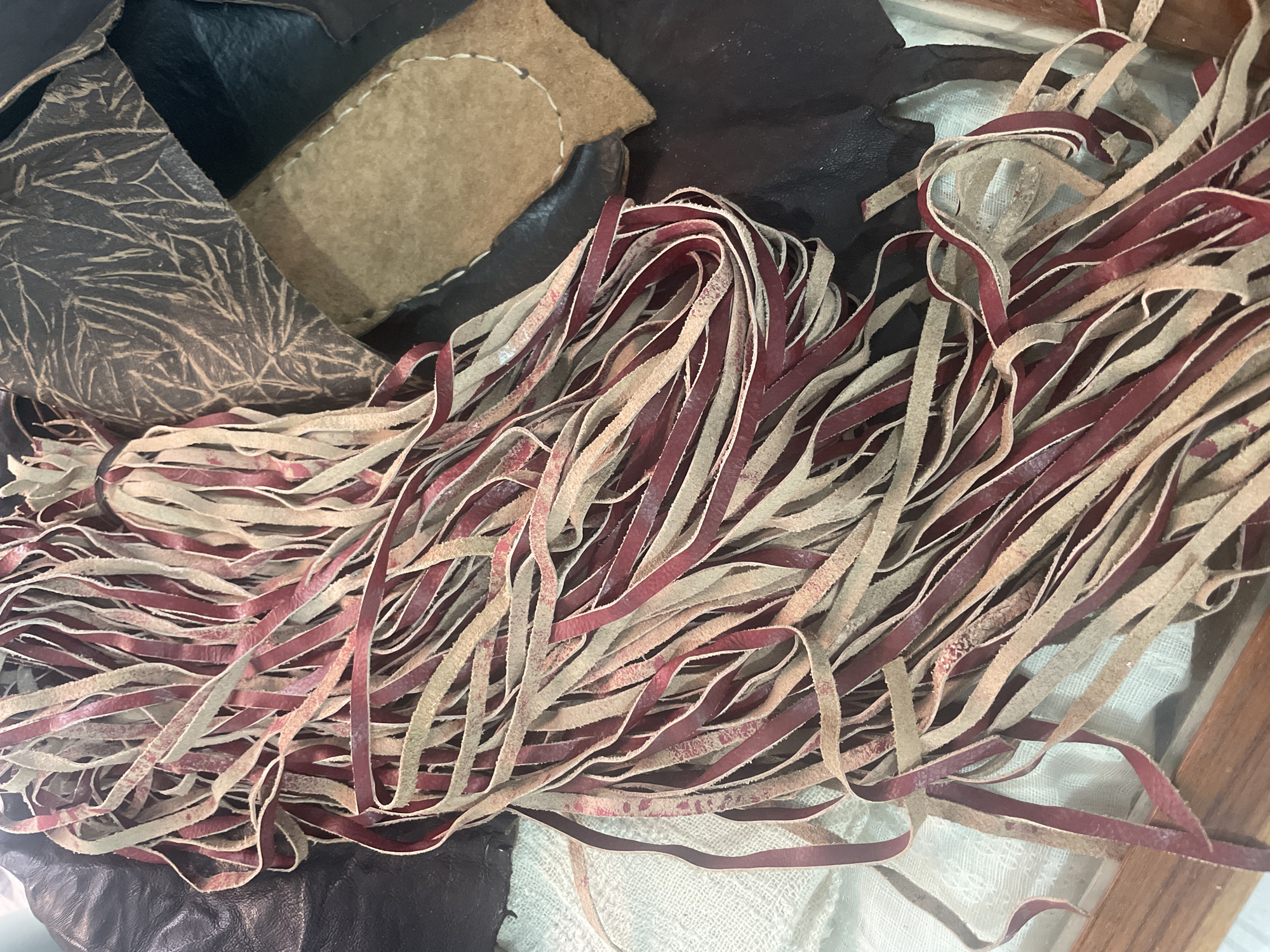 The image shows a close-up view of a pile of leather fringe in various shades of maroon and tan. The fringe is long and thin, with some strands appearing frayed or uneven. The fringe is arranged in a loose pile, with some strands cascading over the edge of a wooden surface. In the background, there are pieces of dark brown and tan leather, one of which has a decorative embossed pattern of leaves. A white, textured fabric is visible beneath the fringe. The lighting appears to be natural, casting shadows and highlighting the textures of the materials. The scene is indoors, possibly in a workshop or a market stall. There are no people or animals visible in the image, and no specific details suggest a location in Bhagsu, India.