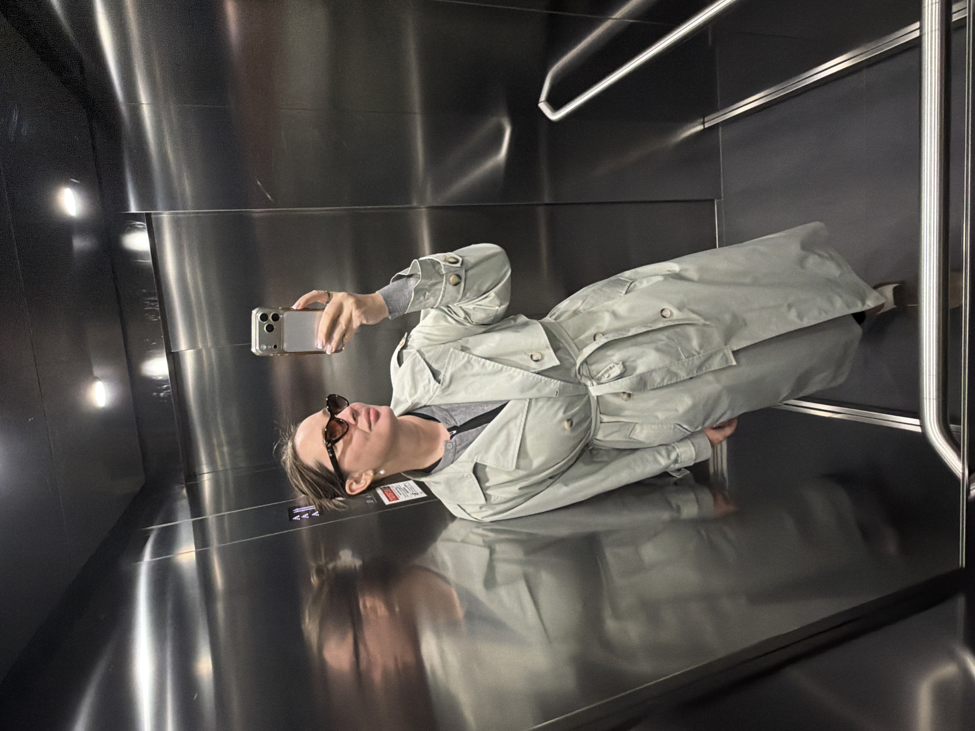 A person wearing a light-colored trench coat and sunglasses is taking a selfie with their phone in an elevator. The elevator has shiny metal walls and handrails. Small lights are visible in the wall behind the person. The person's reflection is visible on the floor. The elevator appears to be located in Moscow, Russia based on the architectural design.
