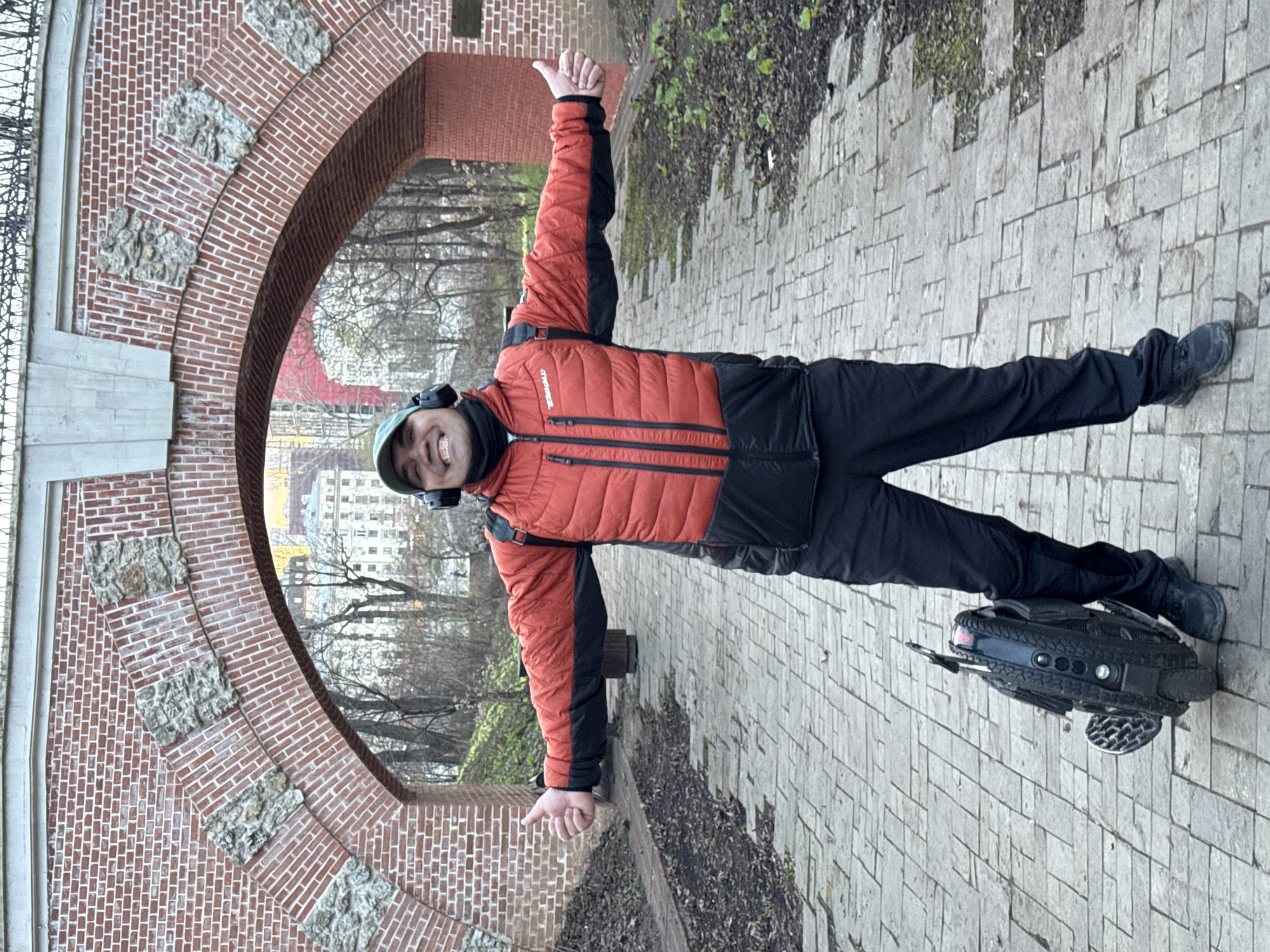 A man stands on an electric unicycle, giving two thumbs up in front of an archway in Moscow, Russia. The man is wearing a green cap, black headphones, a black neck gaiter, an orange and black quilted jacket, and black pants and shoes. The archway is made of red bricks and has gray stone embellishments. Through the archway, a building with a red roof and other buildings can be seen. The ground is paved with gray bricks, and there are trees and some greenery in the background. The unicycle is black with silver footrests.