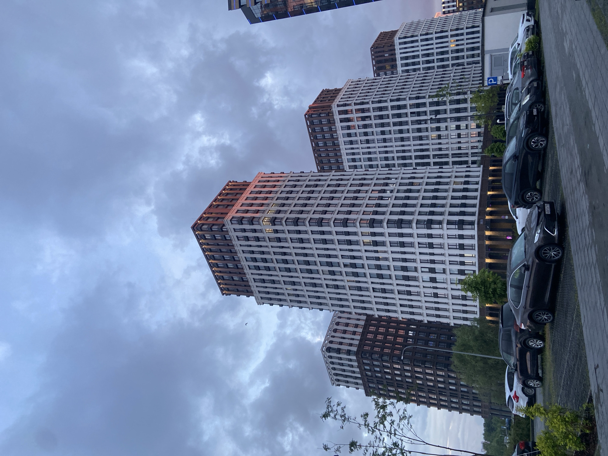 This media file depicts a modern urban scene in Kazan, Russia, likely during twilight or a heavily overcast day. The central focus is a cluster of several contemporary high-rise apartment buildings, characterized by their striking architecture. These buildings feature predominantly white facades with numerous dark-framed windows arranged in a grid pattern. The upper sections and some vertical elements of the buildings are clad in a contrasting brown or bronze material, which catches a soft, warm, possibly golden light, suggesting either the glow of a setting sun or artificial illumination. Several windows across the buildings are lit from within.
In the lower right portion of the image, a row of parked cars is visible on a dark paved surface, bordering a concrete sidewalk. The cars are of various models and colors, mostly dark-toned. A clear blue parking sign with a white 'P' and an arrow is mounted near the parking area. Small trees and sparse foliage line parts of the sidewalk and are scattered around the buildings. One parked car's license plate is partially visible, showing "X809XX 116," with "116" being the regional code for the Republic of Tatarstan, where Kazan is located.
The sky is heavily overcast with thick, grey clouds, contributing to the subdued lighting of the scene. A tiny bird is captured in flight against the cloudy backdrop. The scene conveys a quiet, architectural presence within a modern residential or mixed-use urban environment. No people are visible, and there is no overt activity taking place.