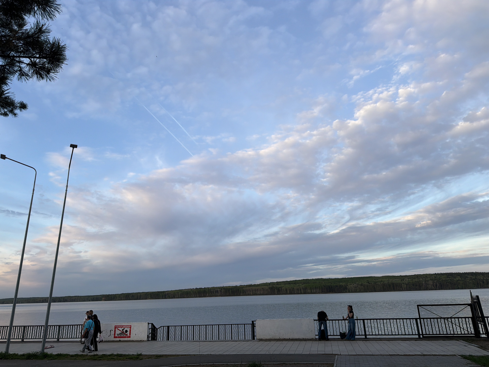 This image depicts a waterfront scene in the city of Chaikovsky, Russia. The sky is partially cloudy with contrails visible. Below the sky is a body of water that appears to be a lake or wide river. A forest line is visible on the opposite bank.
A pedestrian walkway runs along the near side of the water, separated from the water by a black metal fence. Three individuals are walking on the left. A woman stands alone at the fence on the right of the image. A warning sign against swimming is affixed to a white concrete partition along the fence. Streetlights are present near the walking path, and a section of trees is at the top left of the image.