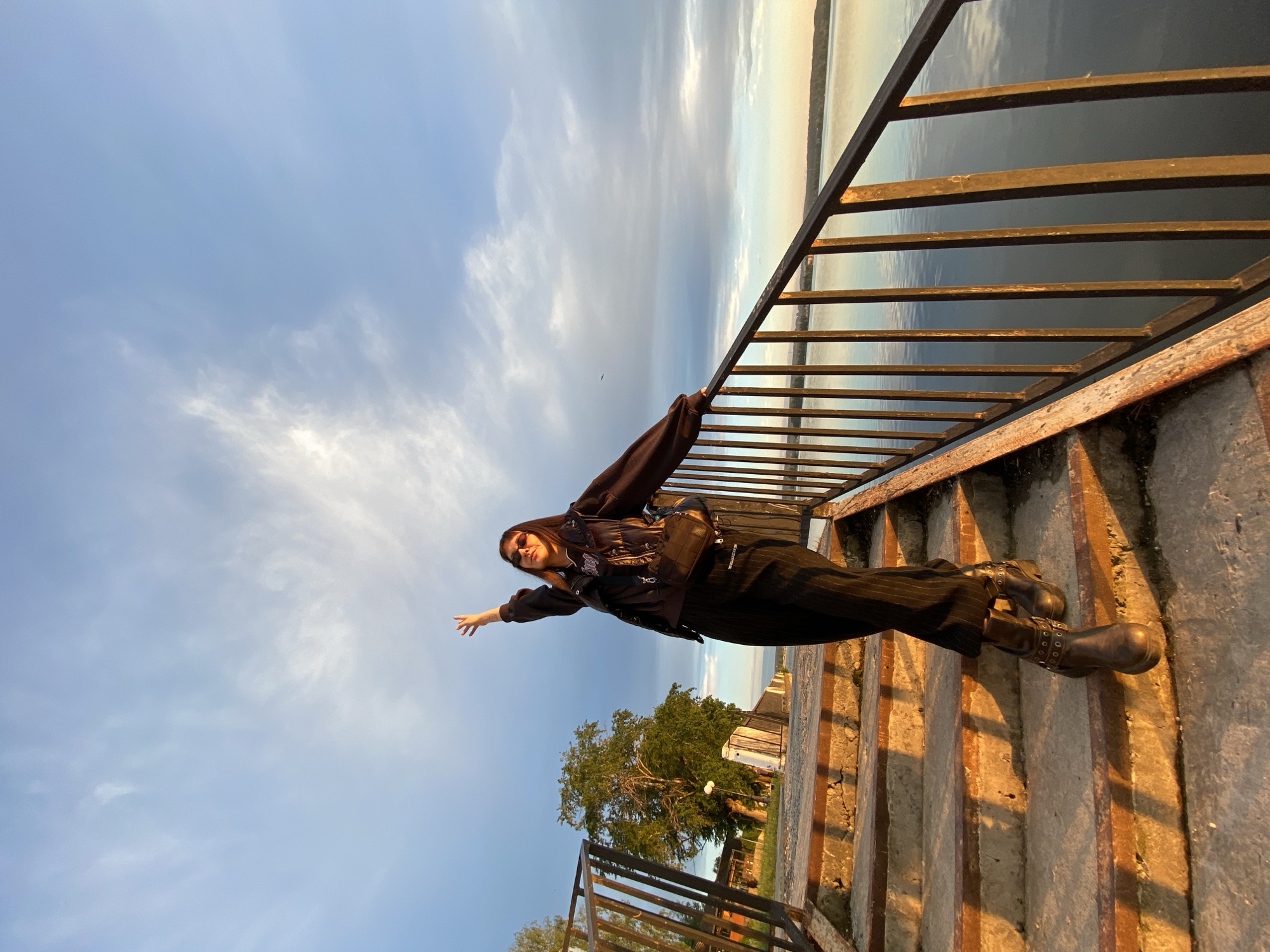 A young woman stands diagonally on concrete stairs with a dark metal railing in Votkinsk, Russia. She wears a dark hooded jacket, black pinstripe pants, and studded boots. She has both arms outstretched, one resting on the railing. The background features a river or large body of water with a cloudy sky and distant shoreline. A building and some trees are partially visible at the bottom of the stairs.