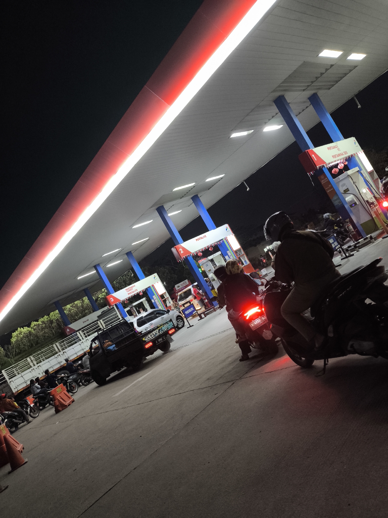 At night, in the city of Waringinkurung, Indonesia, a gas station with a large illuminated red and white canopy is the scene of activity. Multiple vehicles, including cars and motorcycles, are present at the pumps. Several individuals are seen operating or waiting on motorcycles. A black pickup truck is parked at the gas station, and a white truck with a trailer is visible in the background. Traffic cones are placed on the ground. The lighting is predominantly artificial from the station's lights and vehicle headlights and taillights, with a dark sky overhead.
