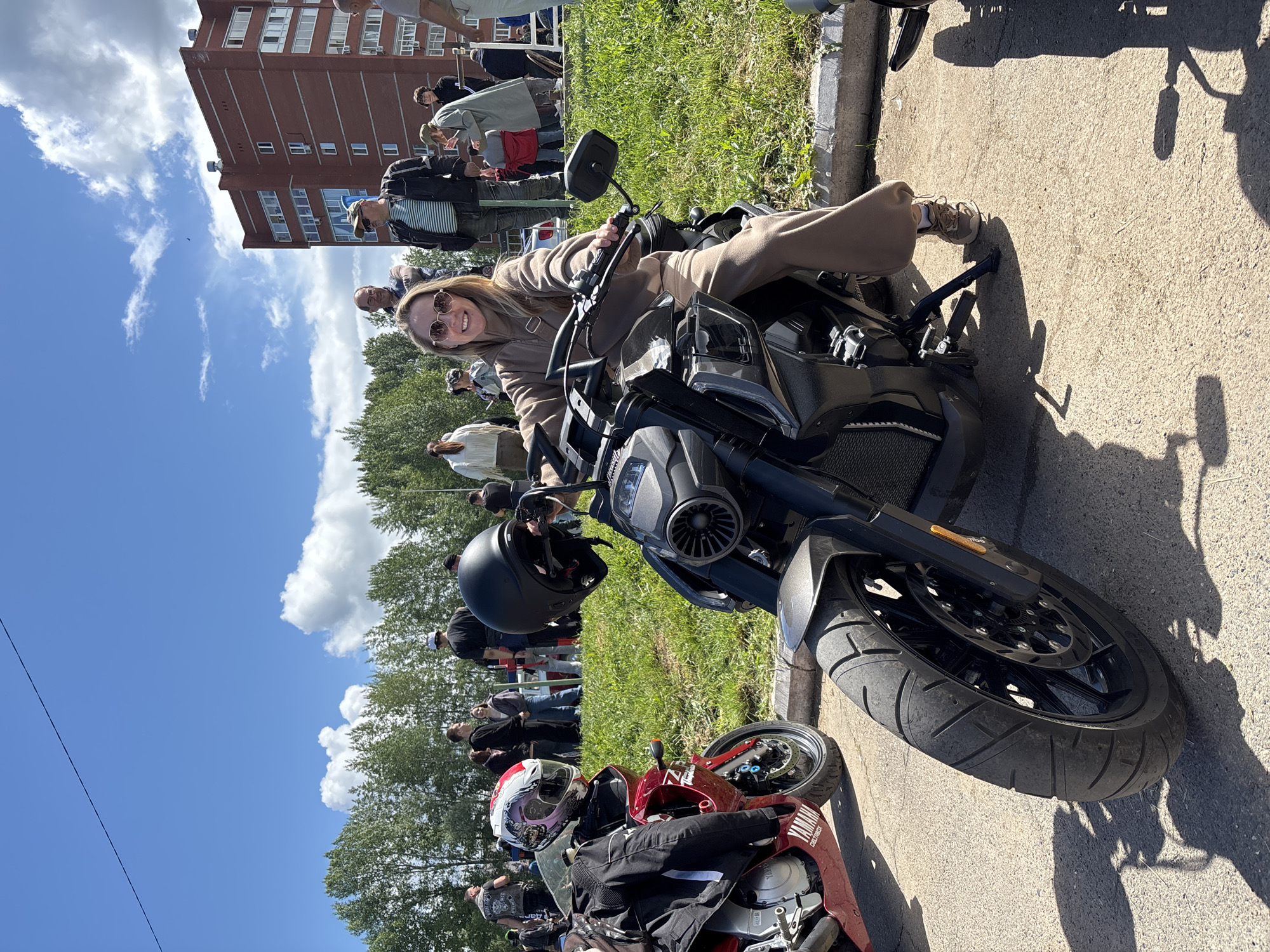 The image shows a woman on a motorcycle. She is fair-skinned, with light brown hair and sunglasses, and wears a tan-colored jumpsuit. She is smiling at the camera. The motorcycle is black and gray, and parked on a paved surface. To the left and behind her are a group of people standing and watching. To her left and forward is another red motorcycle with a person in riding gear. Behind the crowd is a grass area, trees, and a brick apartment building. The sky is blue with some clouds. The location is Chaikovsky, Russia.