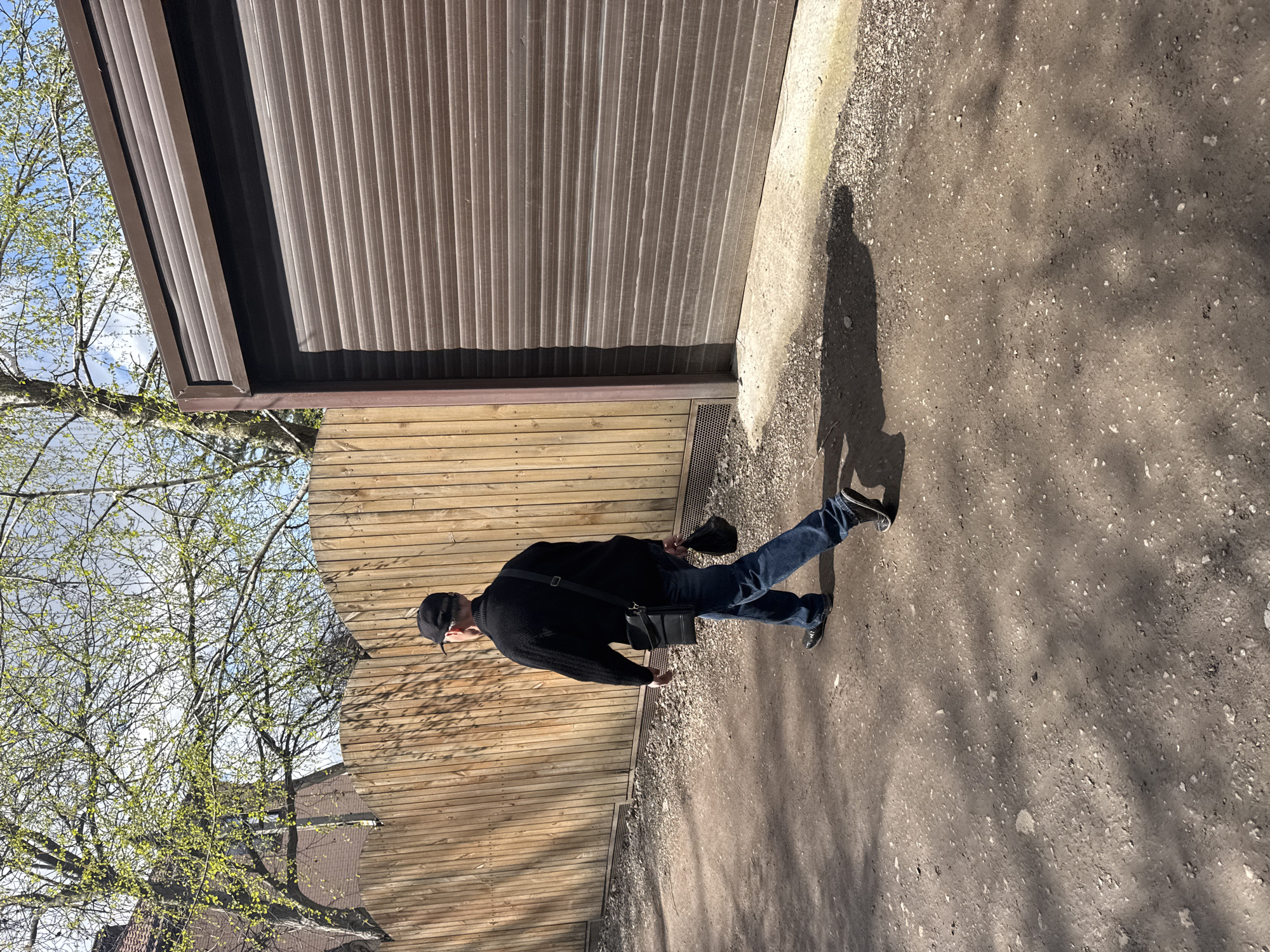 The image shows a man walking on a dirt path next to a building. The building has a brown metal corrugated facade and a wooden fence underneath. The man is wearing a black sweater, blue jeans, and a black baseball cap. He has a black shoulder bag. Trees with new leaves are visible on the left side of the image. The location is likely in Rostov-on-Don, Russia.