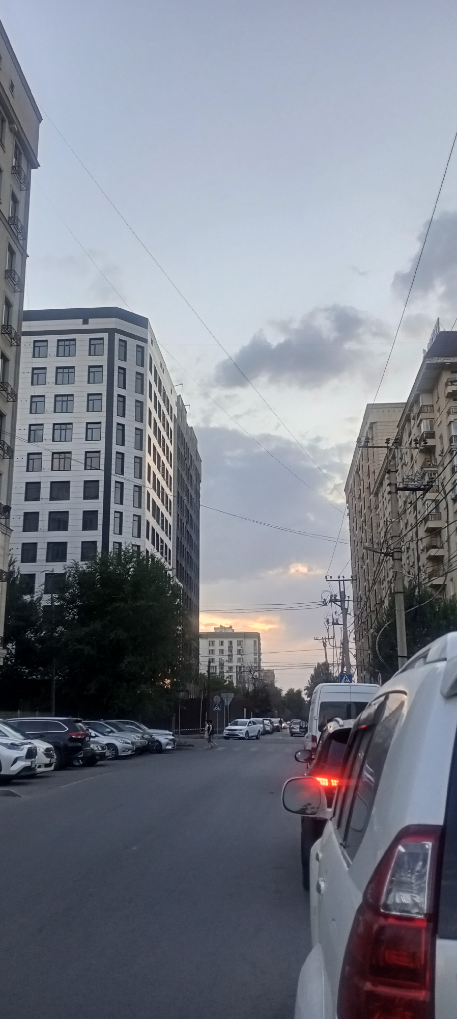 The image shows a street scene in Bishkek City, Kyrgyzstan, during what appears to be late afternoon or early evening. Tall, modern buildings with many windows line both sides of the road. The sky is overcast with patches of lighter clouds, and a hint of sunset can be seen in the distance. Cars are parked along the left side of the street, and a line of traffic is moving down the road, with the back of a white SUV prominently in the foreground on the right. A few individuals are visible walking on the sidewalk and crossing the street. Electrical wires are strung across the sky. The overall impression is of a typical urban environment with some traffic congestion.
