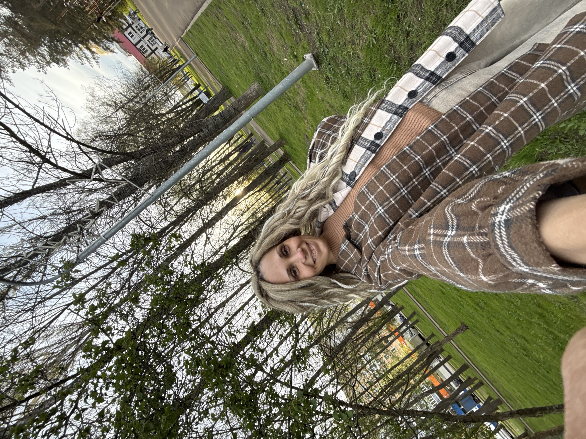 The image shows a blonde woman with a brown and white plaid shirt in a grassy area of what appears to be a park. The location is identified as Rusanovo, Russia, as specified in the query. Trees with bare branches fill the background, along with residential buildings. The woman is smiling at the camera.
