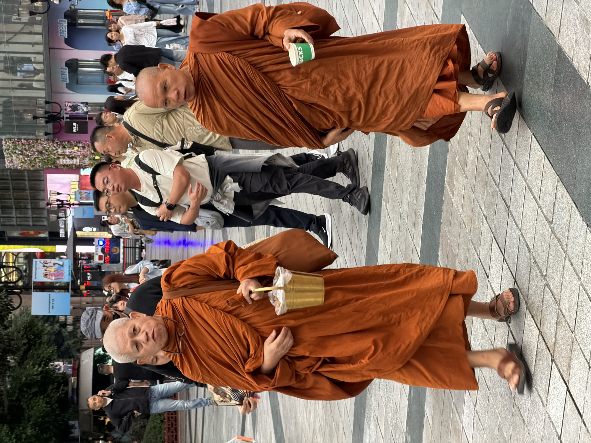 In Yuzhong District, China, two Buddhist monks in saffron robes walk across a paved outdoor plaza. The monk on the left is older with a shaved head and is carrying a gold bucket with a plastic bag inside. The monk on the right also has a shaved head and is holding a green Starbucks cup. They are walking amongst a crowd of people, some of whom are dressed in casual modern clothing. The environment includes storefronts and signage in the background, suggesting a commercial area.