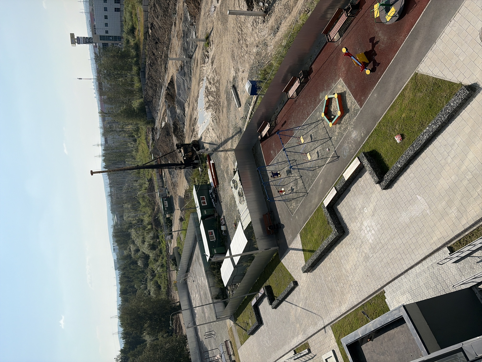 The image depicts a construction site adjacent to a newly developed residential area in Yekaterinburg, Russia. In the foreground, a paved courtyard area features a playground with swings, a sandbox, a seesaw, and a merry-go-round. Benches are strategically placed around the playground, and small grassy areas with gabion retaining walls are interspersed throughout the courtyard. A modern building with dark grey accents and balconies is partially visible on the left.
The construction site itself is a large, excavated area with piles of dirt and gravel. A pile driver machine is positioned in the middle of the site, indicating ongoing foundation work. Several temporary green office containers are situated along the perimeter fence of the construction zone. In the background, a dense line of trees separates the construction site from a distant urban landscape featuring industrial buildings and power lines. The sky is clear and blue, suggesting a sunny day.