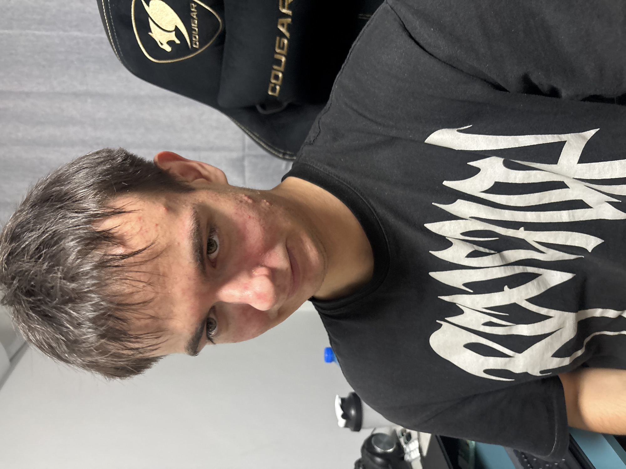 A young man is seated in a black and gold Cougar gaming chair. He is wearing a black t-shirt with a large white graphic. The background consists of a gray wall. A water bottle and some other objects are visible on a desk in the lower part of the frame. The picture appears to have been taken indoors. There is no indication that this image was taken in Plovdiv, Bulgaria.
