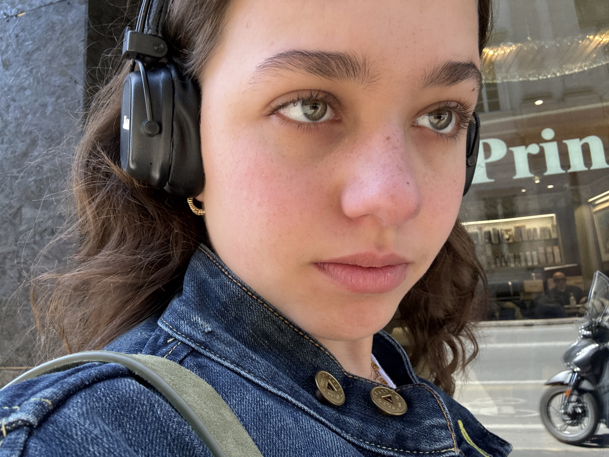 A young woman wearing black headphones and a denim jacket is standing outdoors. The environment is identified as Milan, Italy, indicated by the reflection of a store with "Prin" visible in the store's window. The woman's eyes are hazel. The background includes a parked motorcycle and a person sitting inside the store. The denim jacket is fastened with buttons marked "G-Star." The woman has an earring in her left ear. She carries a bag with a green strap.