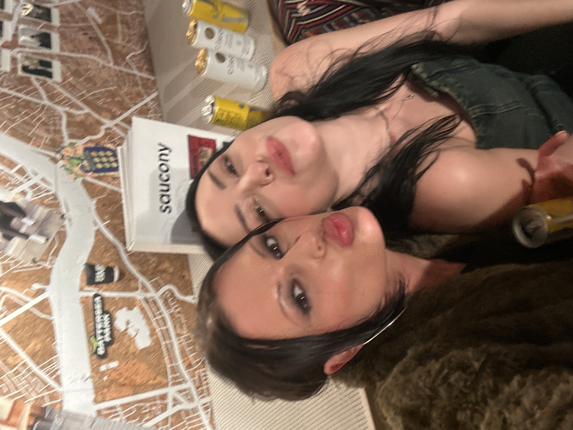 The image displays two adult females posing closely for a selfie-style photograph in an indoor commercial or social establishment. The background wall features a prominent three-dimensional map of London, specifically highlighting "BATTERSEA PARK" and indicating the location is in or near Battersea, London, United Kingdom.
The female on the left has dark hair, dark eye makeup, large silver hoop earrings, and is making a "kissy face" gesture. She wears a dark, textured, possibly faux fur, jacket. The female on the right has long dark hair, a visible nose ring, and a slight smile. She wears a dark denim-like strapless top and a thin silver necklace.
On a surface immediately behind and to the right of the subjects, a white display stand or box is visible, featuring the brand name "saucony." To the right of this, four beverage cans are arranged: two yellow cans, one fully visible, and two white cans with gold accents. A partial view of a hand holding a can or bottle is present in the lower center.