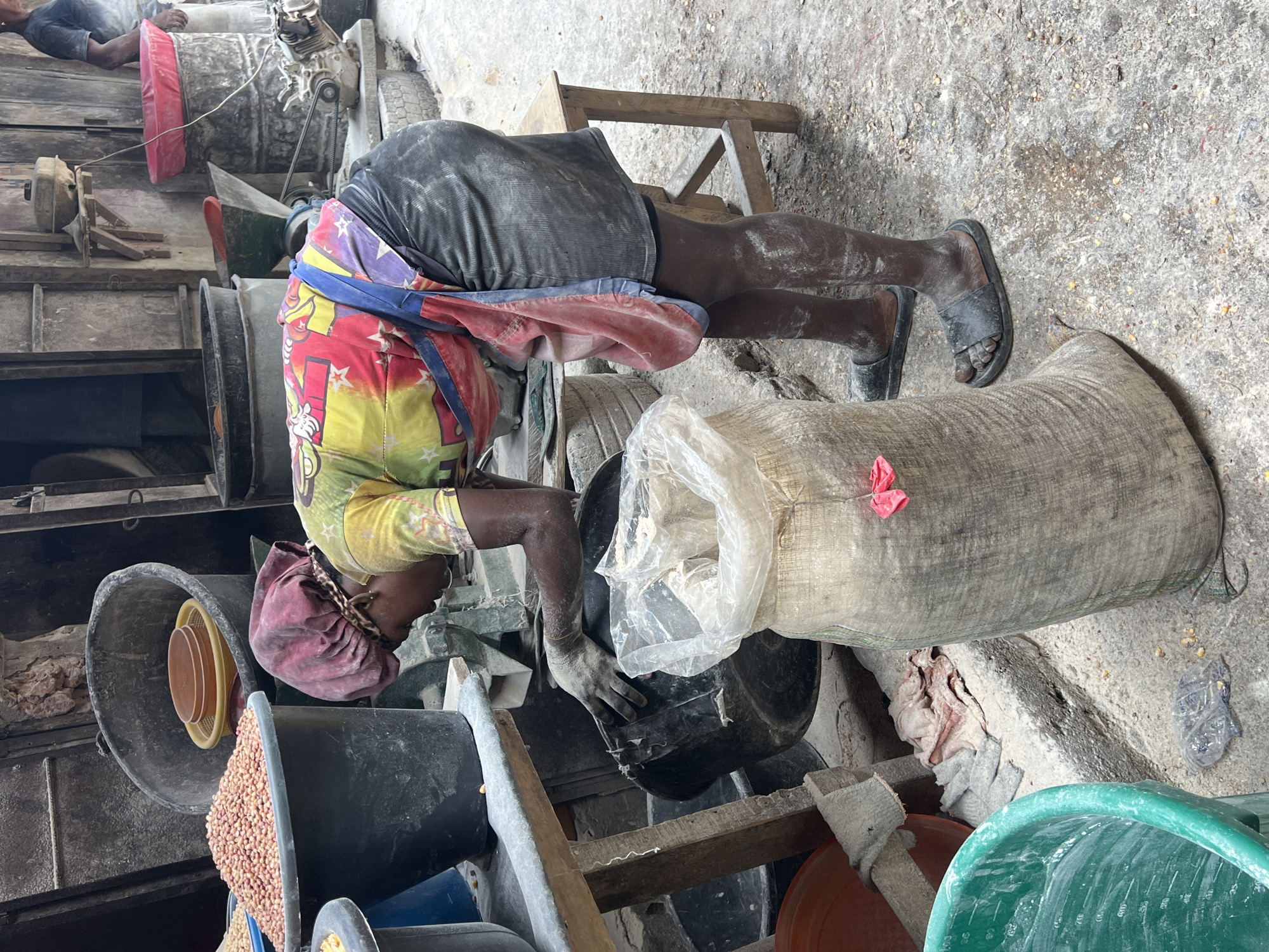 An individual is shown working in a dusty environment, presumably a processing or storage area. The person is bent over, their hands near a black container and a large sack filled with what appears to be a granular substance, possibly grains. The environment is cluttered with various buckets, barrels, and other containers, along with machinery or equipment parts in the background. The scene is grimy and suggests an industrial or agricultural setting. Location information is not possible to determine.