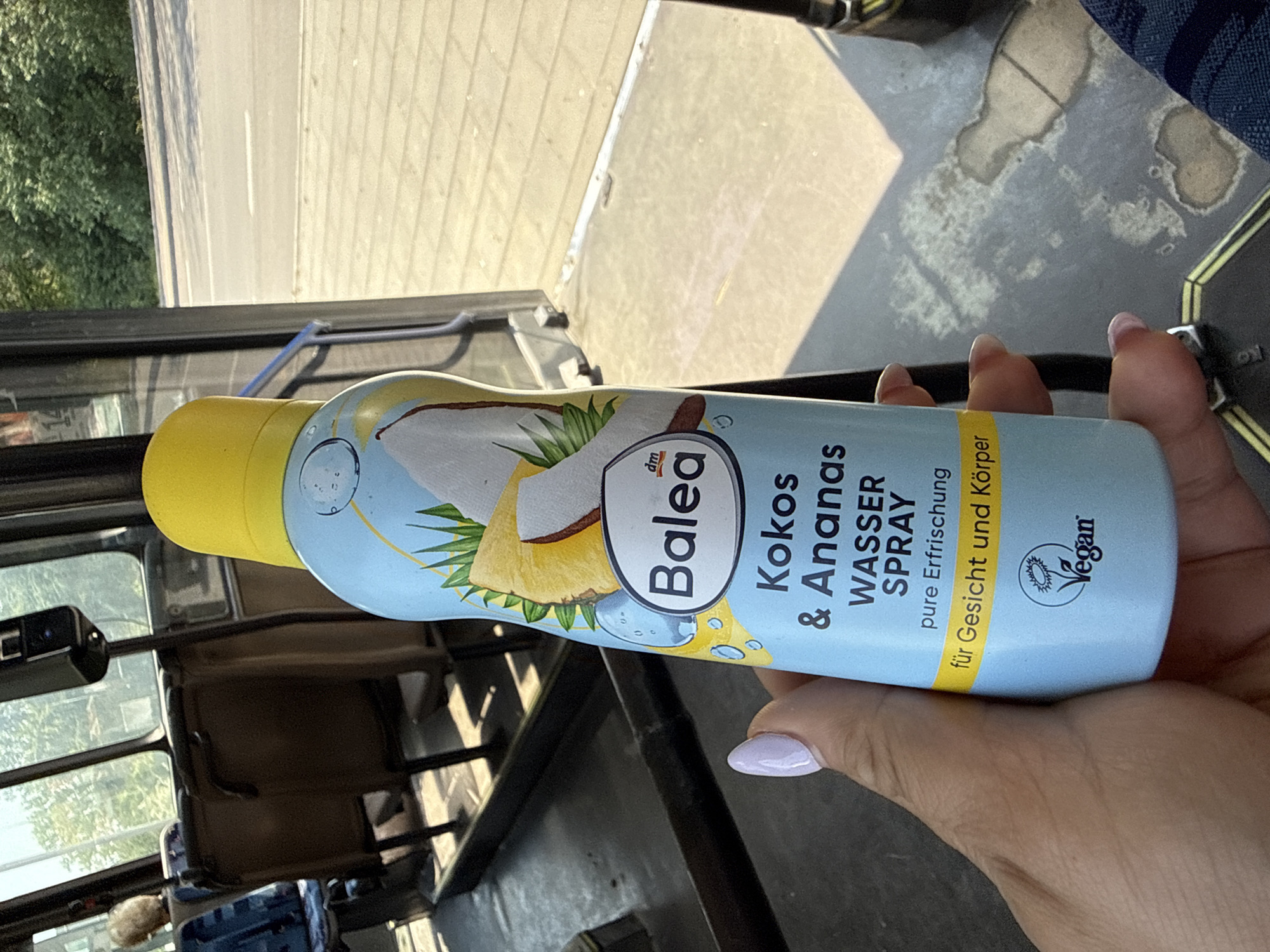 The image shows a person's hand holding a light blue spray bottle inside what appears to be a moving vehicle, likely a bus or car, during daytime. The hand, with light purple nail polish, holds the bottle vertically in the center-right of the frame.
The bottle is a "Balea Kokos & Ananas WASSER SPRAY" (Coconut & Pineapple Water Spray) from the brand "dm Balea". It has a yellow cap and features illustrations of coconut and pineapple. The text on the bottle also reads "pure Erfrischung" (pure refreshment) and "für Gesicht und Körper" (for face and body). A "Vegan" logo is visible near the bottom right of the label.
The background reveals the vehicle's interior, with dark dashboard elements and window frames. Through the window, outside scenery is visible, suggesting the vehicle is on a road. On the left side, lush green foliage (trees or bushes) can be seen. On the right, there's a bright, light-colored surface, possibly a road or pavement, with some lines or textures. Strong sunlight casts clear shadows, indicating a bright, sunny day. A small, black device, possibly a dashcam, is mounted on the dashboard on the left. The overall setting is consistent with someone traveling and using a personal care product.