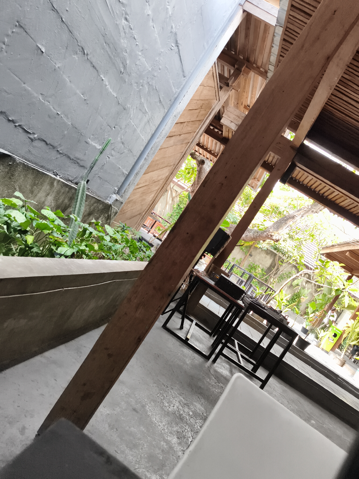 This image captures a portion of an outdoor seating area in Nologaten, Indonesia. The scene is dominated by an angled, weathered wooden beam running diagonally across the frame. To the left, a rough, grey concrete wall with a textured surface is visible, with a planter box containing lush green foliage and a tall, slender cactus. Beyond the planter, a grey pipe runs along the top of the wall. The background reveals a wooden roof structure with exposed beams and rafters. In the mid-ground, several dark metal tables with dark tabletops are arranged, suggesting a cafe or restaurant setting. Further in the background, there is dense green vegetation and what appears to be another building or structure. The ground is a grey, concrete surface.