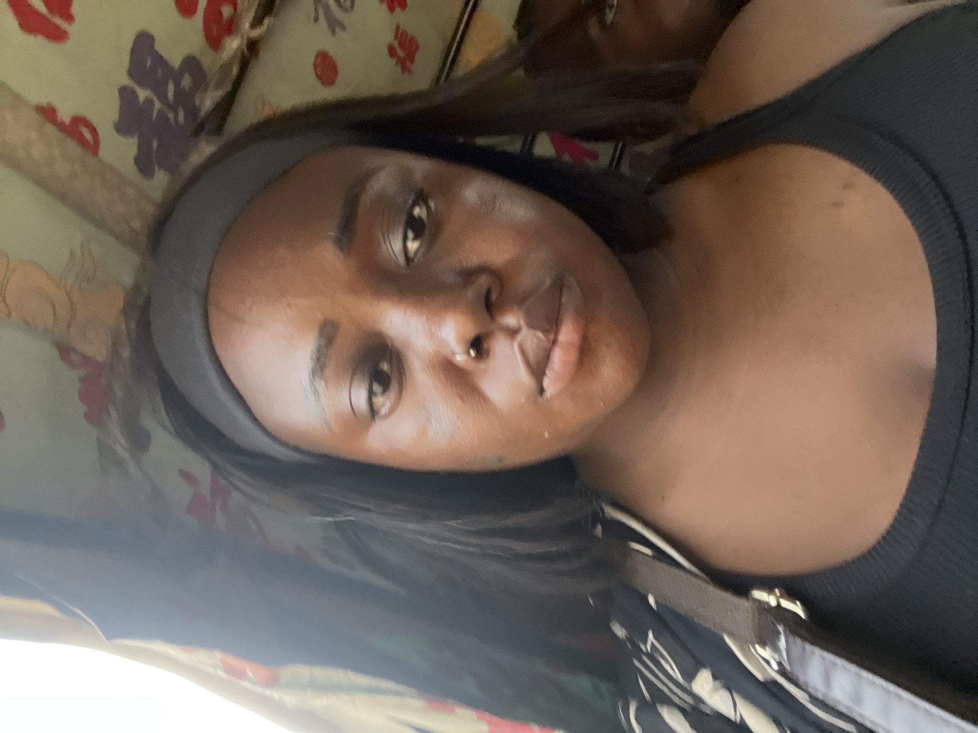 The media file is a close-up, portrait-style photograph of a young Black woman. She is wearing a black tank top and a dark headband. A portion of a patterned bag or garment is visible on her shoulder, featuring black and white abstract designs. The background appears to be a colorful, patterned fabric, possibly upholstery or a decorative cloth, with red and purple motifs. The lighting suggests it is daytime, with natural light illuminating her face. Her expression is neutral or slightly pensive. A nose ring is visible on her right nostril. The image is oriented vertically. There is no visible text in the image.