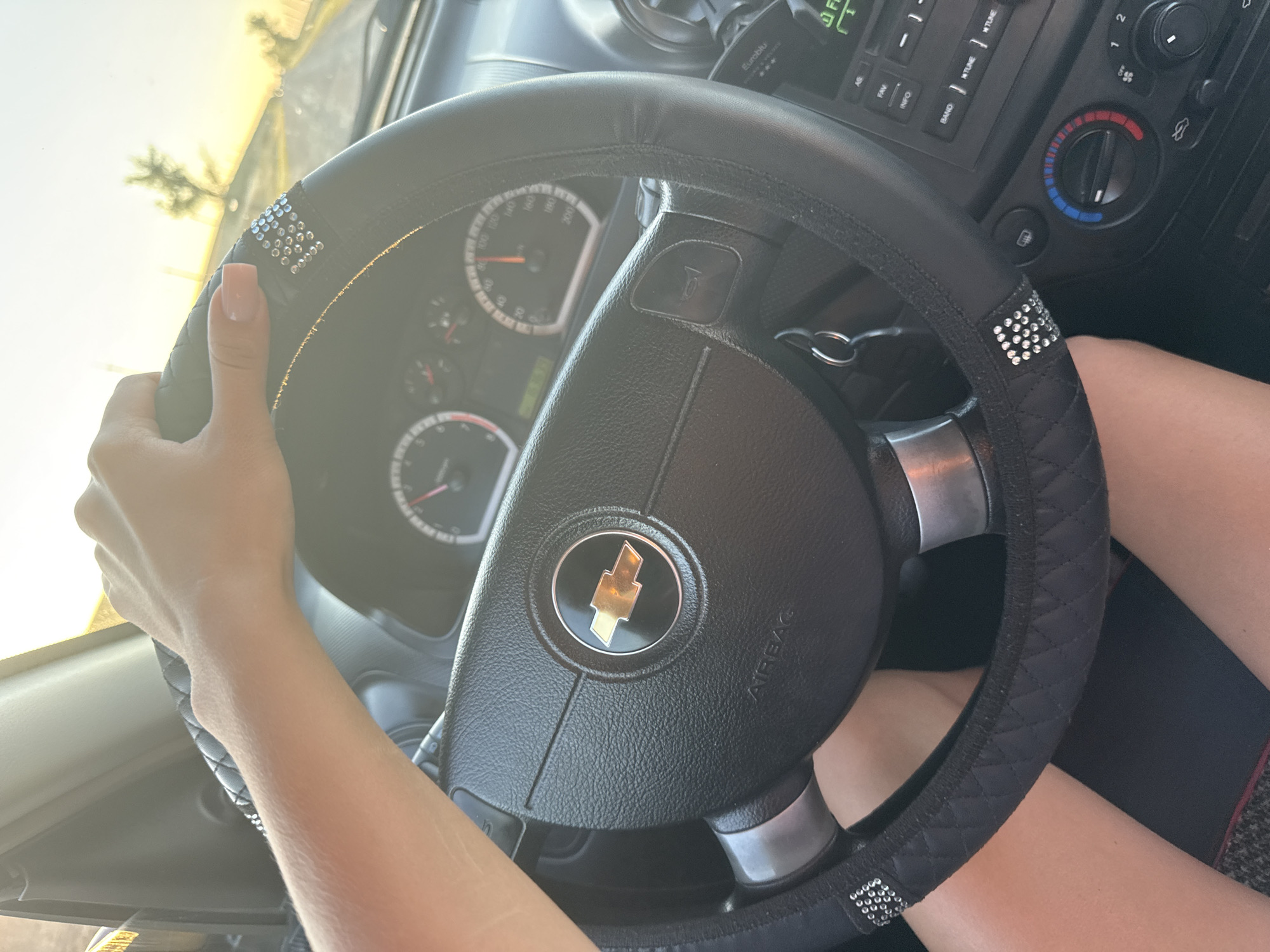 A close-up, eye-level shot shows a person's right hand resting on the steering wheel of a car. The steering wheel is black with a diamond-stitched cover and adorned with small, clear rhinestones. The Chevrolet logo is visible in the center of the steering wheel. The car's dashboard instruments, including a speedometer, are visible in the background. The sun is setting, casting a warm glow and visible through the windshield, creating a slightly overexposed effect. The environment outside the car appears to be a rural or suburban area, with a tree and telephone pole visible in the distance. The image was captured in the city of Iordanovca, Moldova.