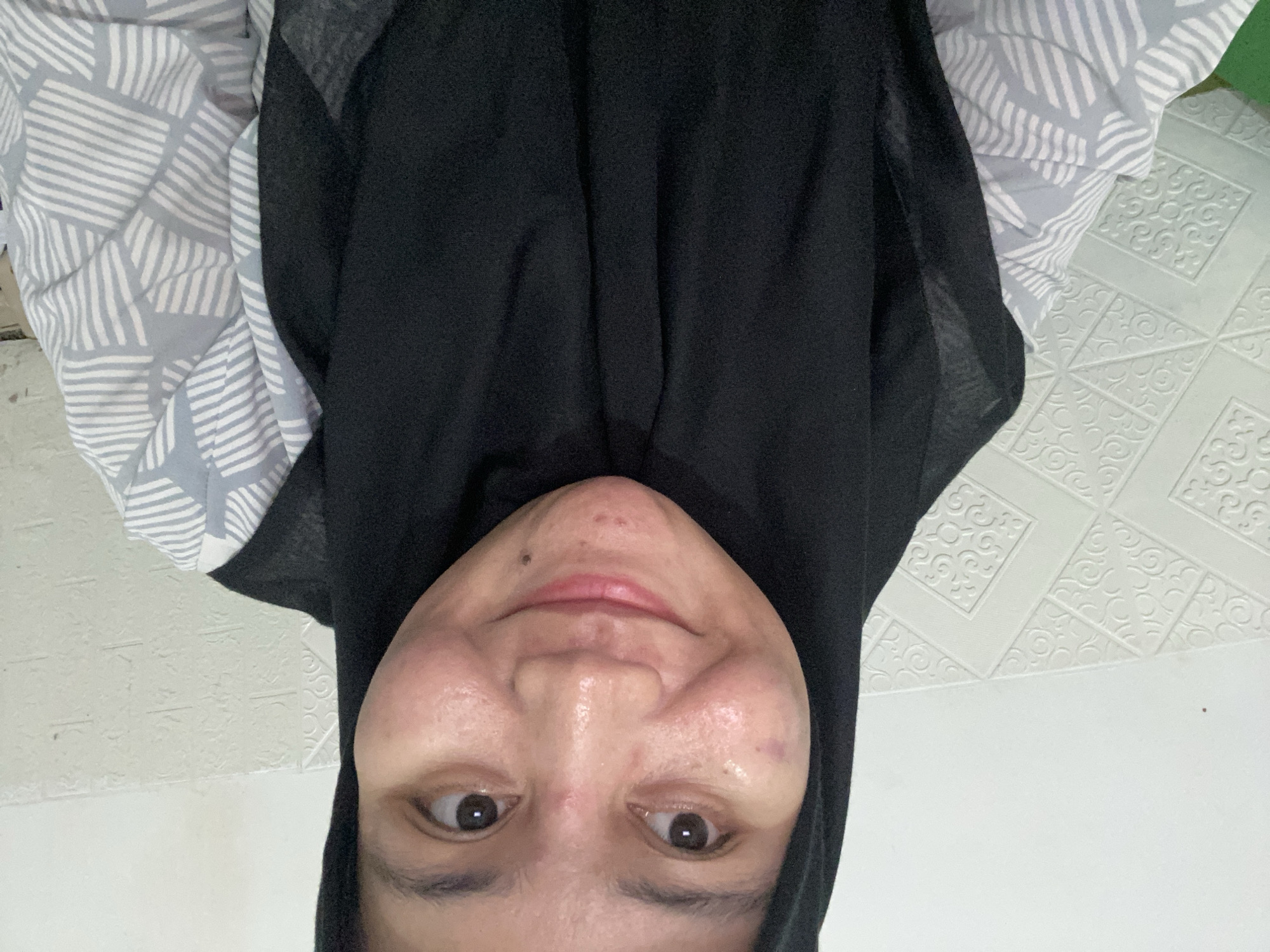 A woman wearing a black hijab and a gray and white patterned shirt smiles at the camera. She is positioned in front of a white wall with a tiled pattern. The woman has acne on her face. The image appears to be a selfie taken indoors, possibly in a residential setting. There are no identifiable landmarks or contextual clues in the image to determine the specific location in Banjarbaru, Indonesia.